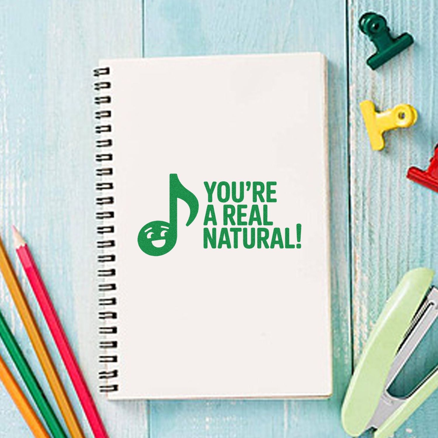 Green stamp seal on white spiral notebook reading Youre a real natural! with colorful stationery on blue wood.