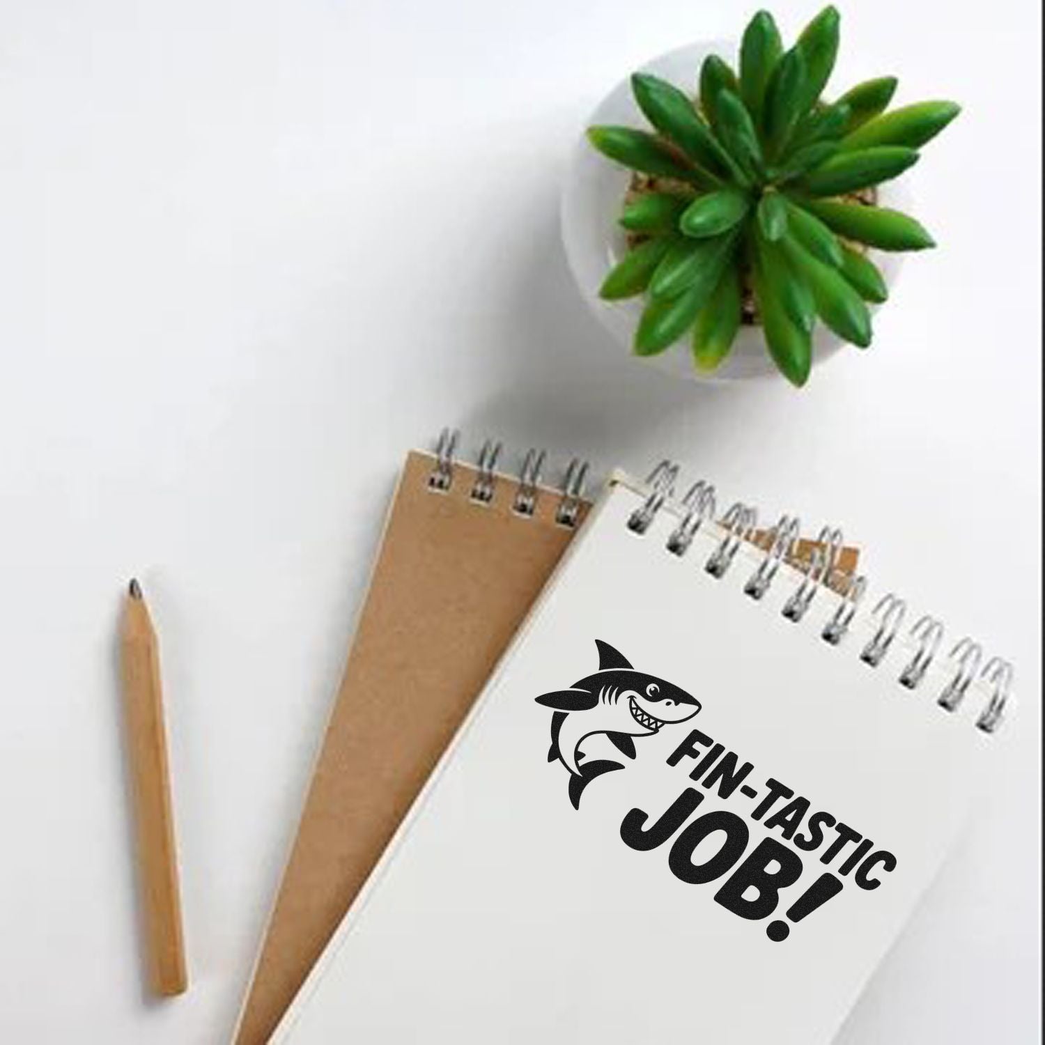 White spiral notebook stamped FIN-TASTIC JOB! with shark graphic, plus pencil, kraft notebook, and green succulent.