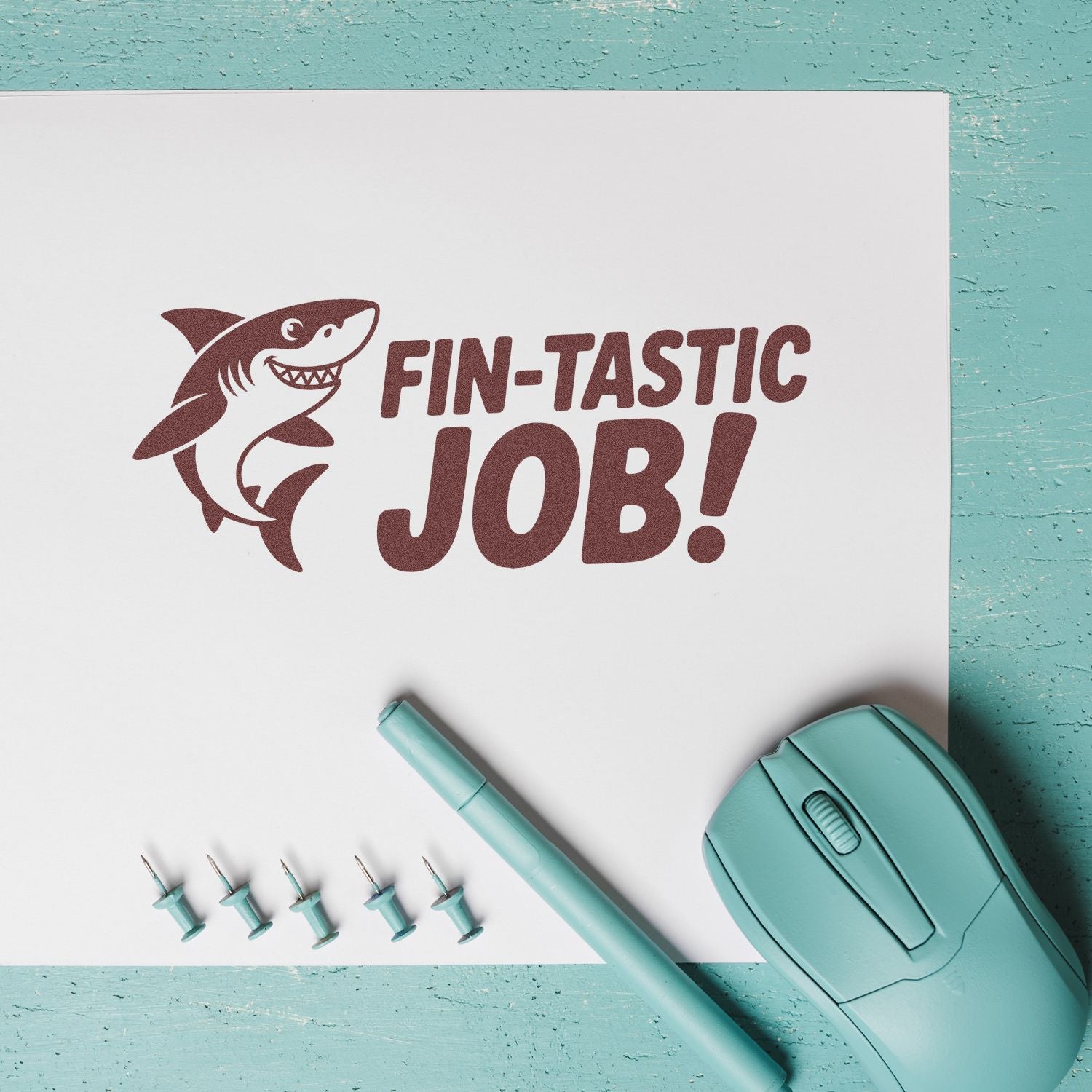 Brown shark FIN-TASTIC JOB! stamp with turquoise seal stamper, pen, and pins on white paper over teal background.
