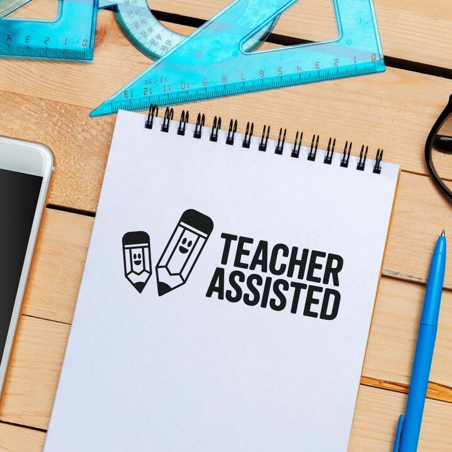 Teacher Assisted stamp/seal with black pencil icons on a white spiral notebook, blue rulers and pen on a wooden desk.