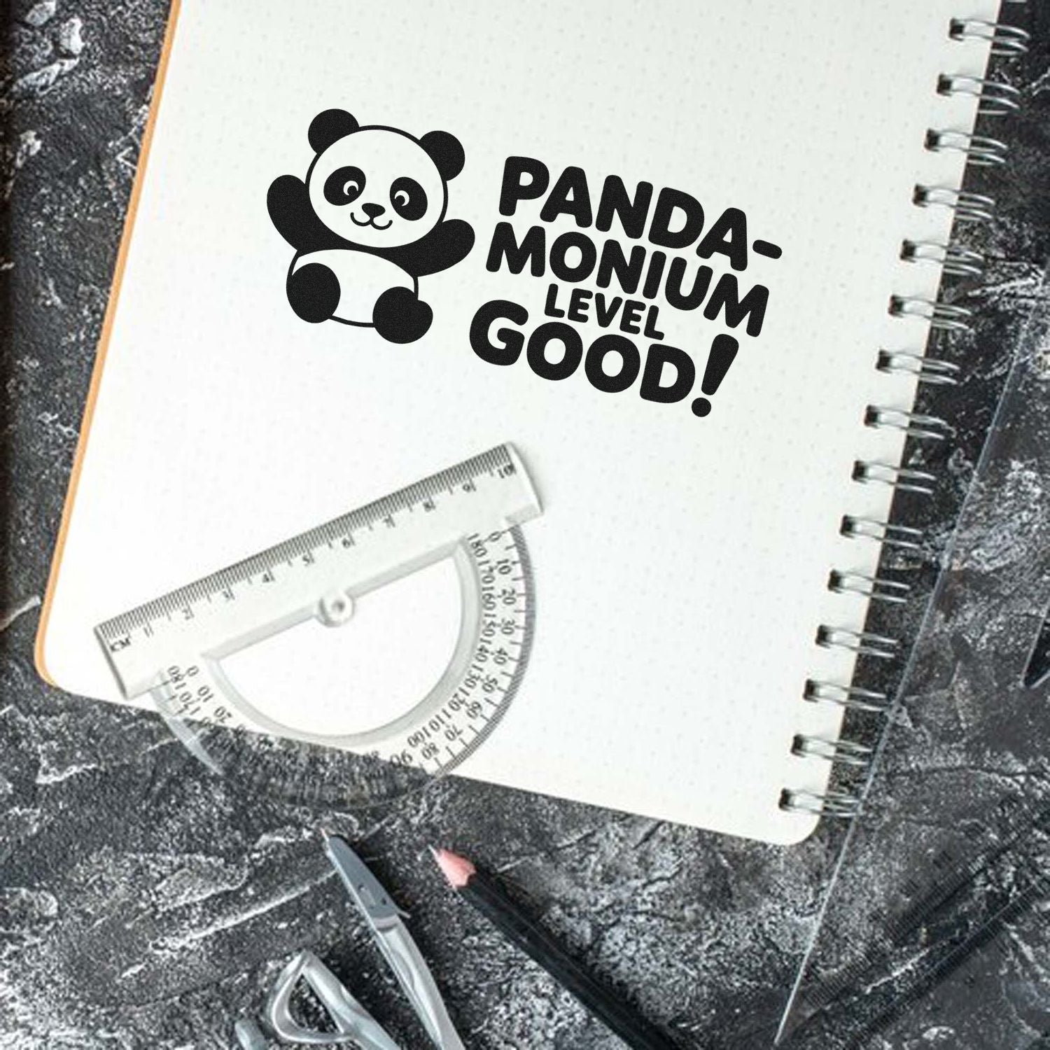 Black panda stamp and seal with PANDA-MONIUM LEVEL GOOD! on a white spiral notebook, plus a clear ruler and protractor.