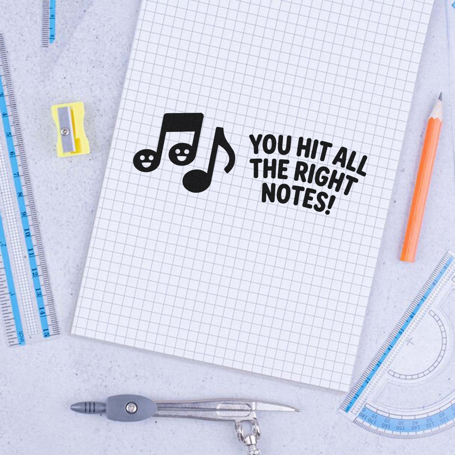 Black music-note stamp and seal with You Hit All the Right Notes! on graph paper, with blue rulers, pencil, and compass.