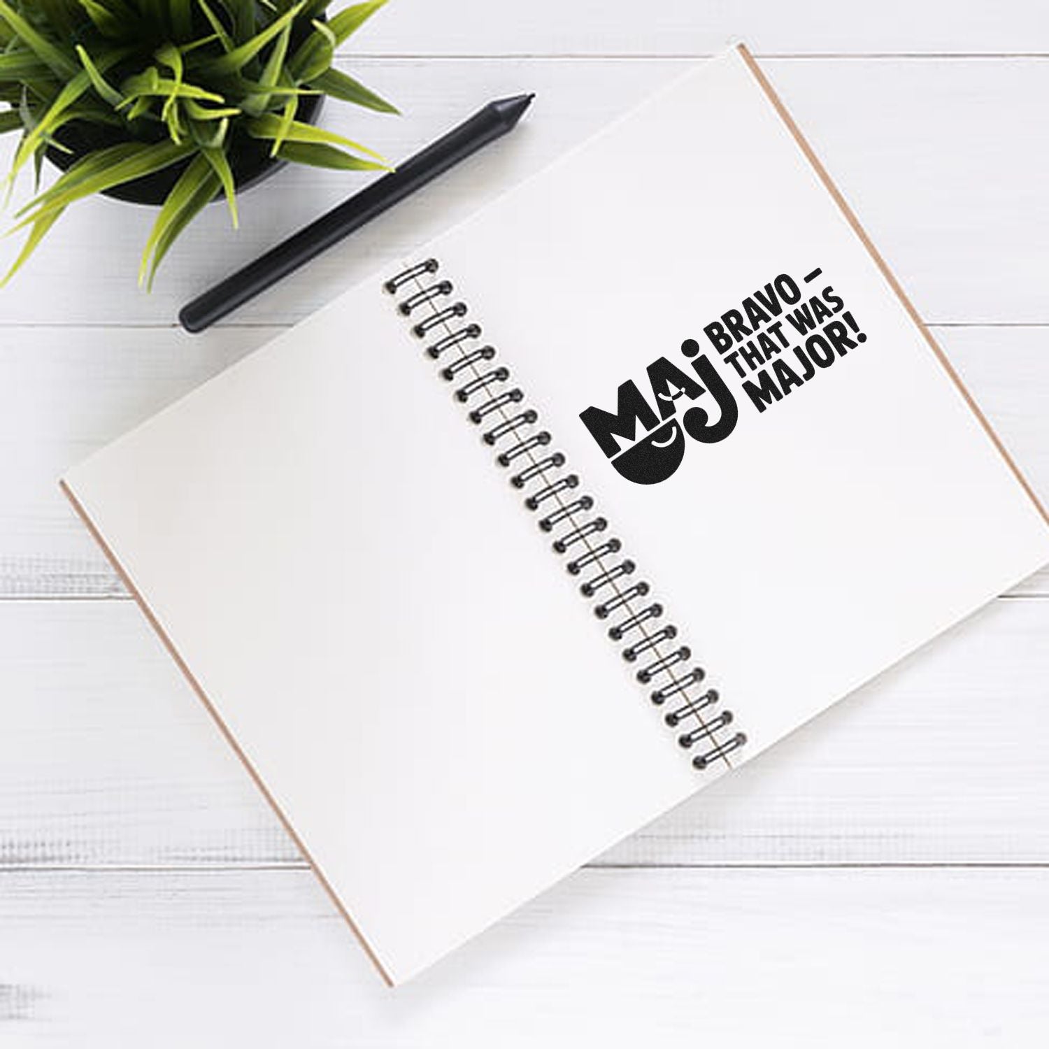 White spiral notebook with black MAJ Bravo stamp seal, black pen, and green plant on a white desk.