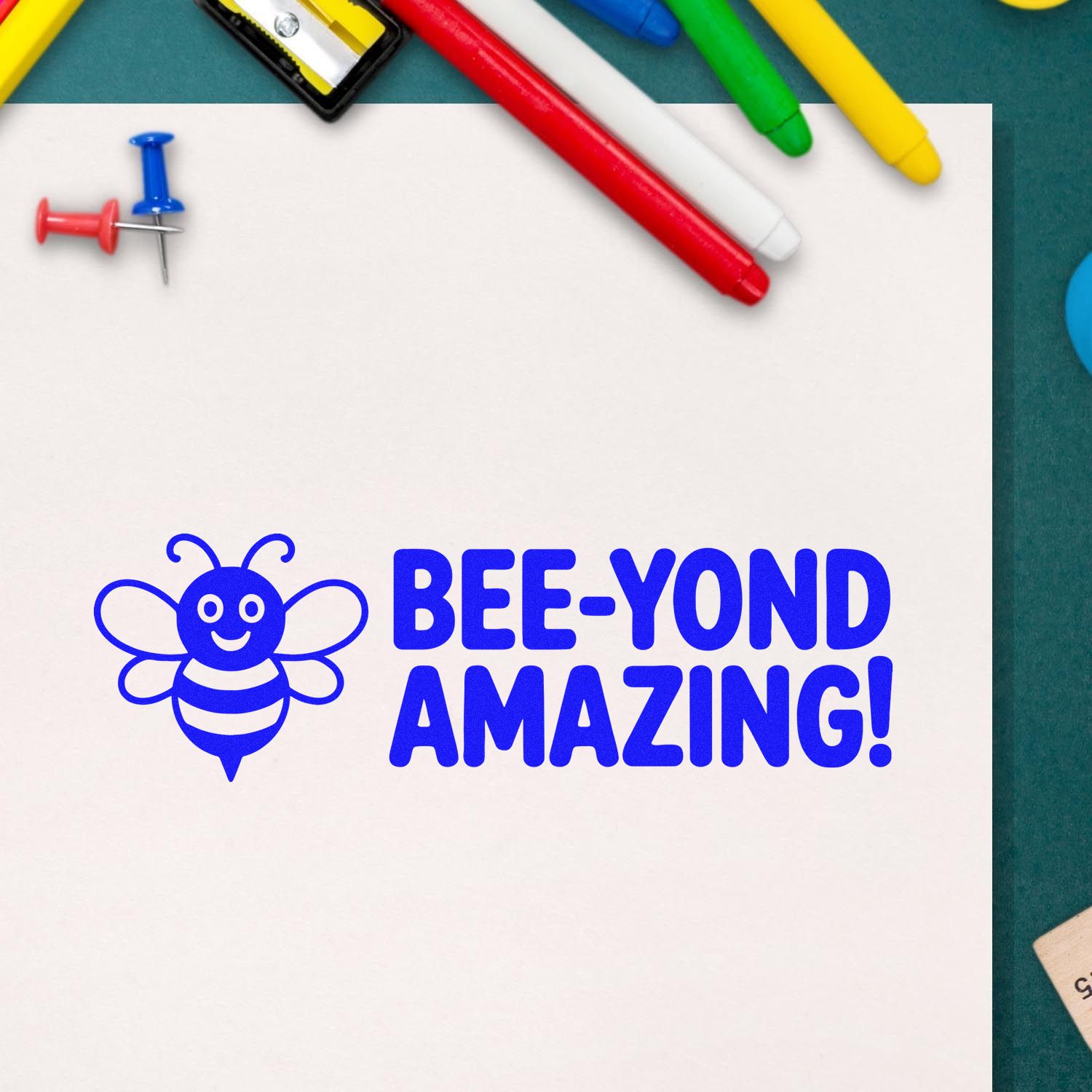 Blue bee stamp seal reading BEE-YOND AMAZING! on white paper, with colorful pens and push pins.