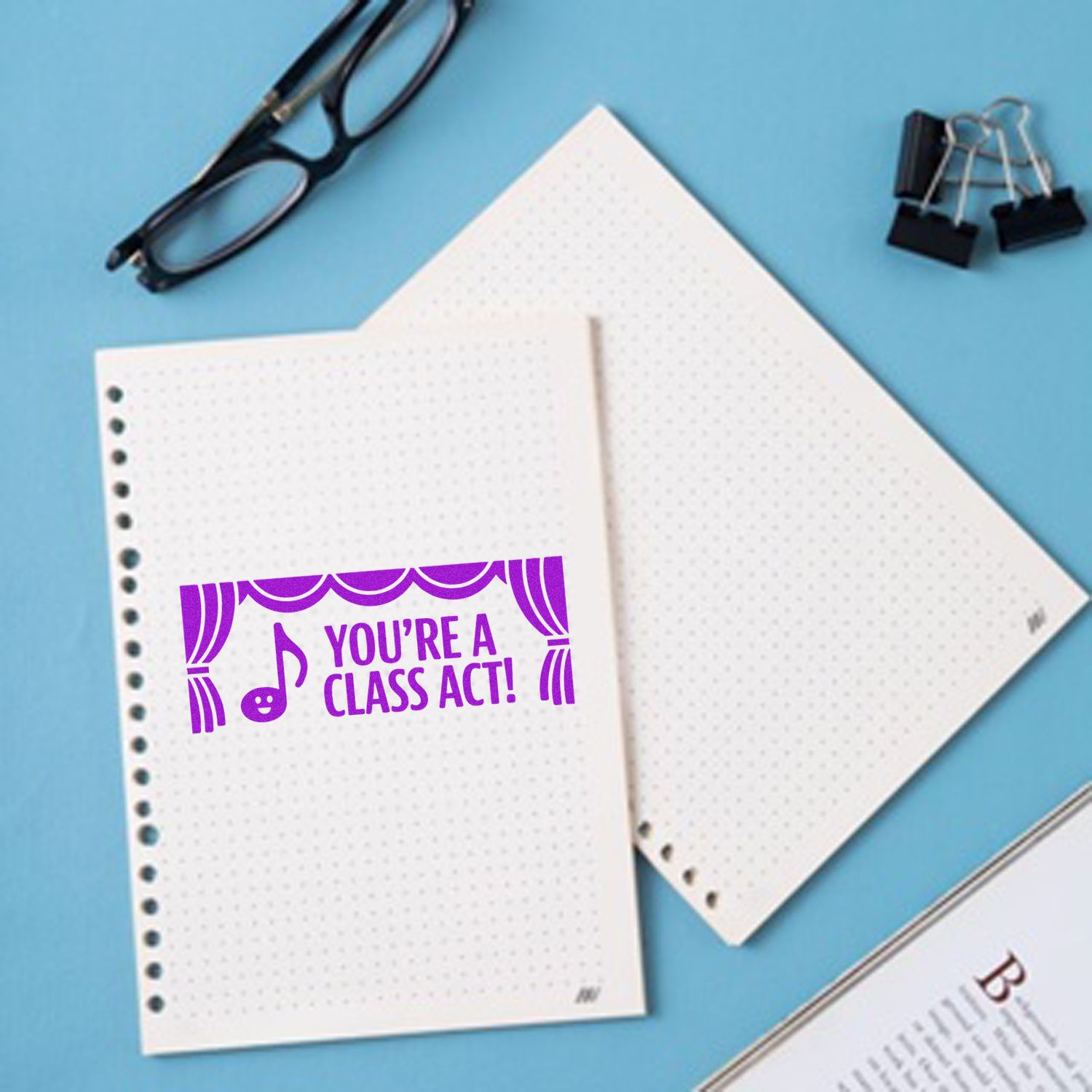 Purple classroom stamp/seal reading Youre a Class Act! on white dotted notebook pages, with black glasses and binder clips on blue.