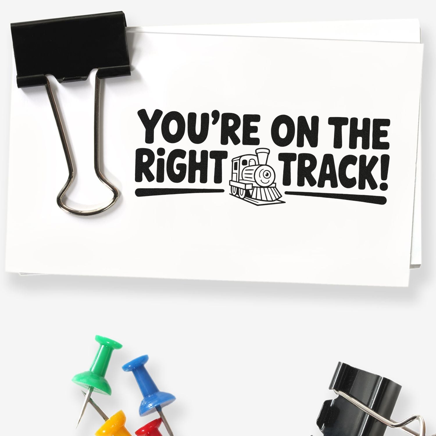 Black binder clip holds white custom stamp sheet reading Youre on the right track! with train icon and colorful push pins.