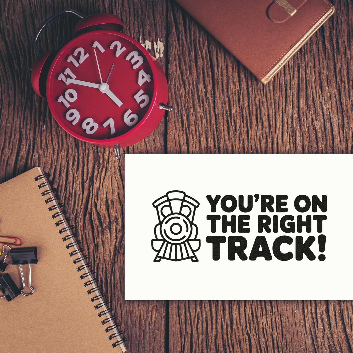 Black train stamp and seal reading Youre on the Right Track! beside a red alarm clock on a wood desk.