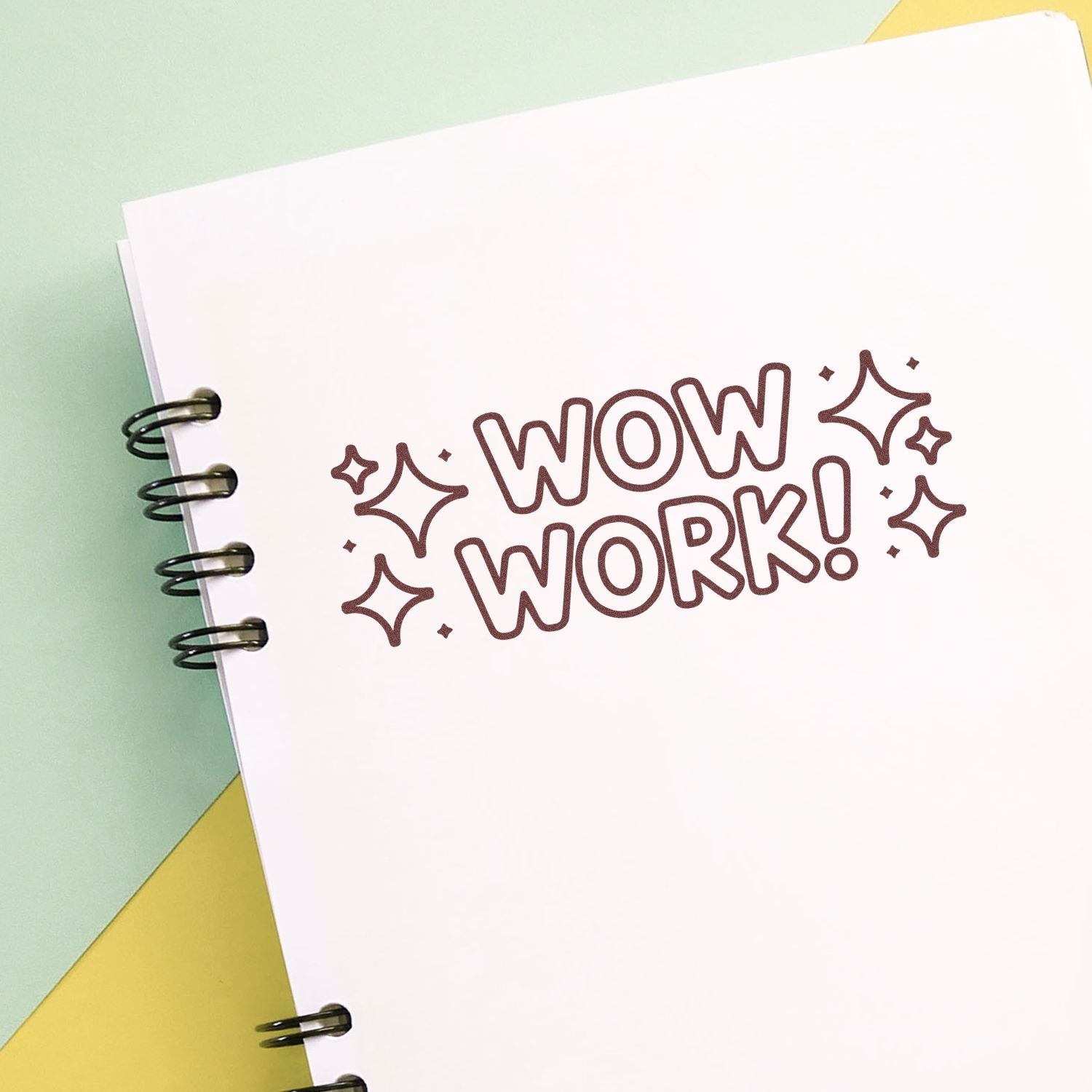 Brown WOW WORK! decorative stamp with sparkles on a white spiral notebook page, against a pastel green and yellow background.