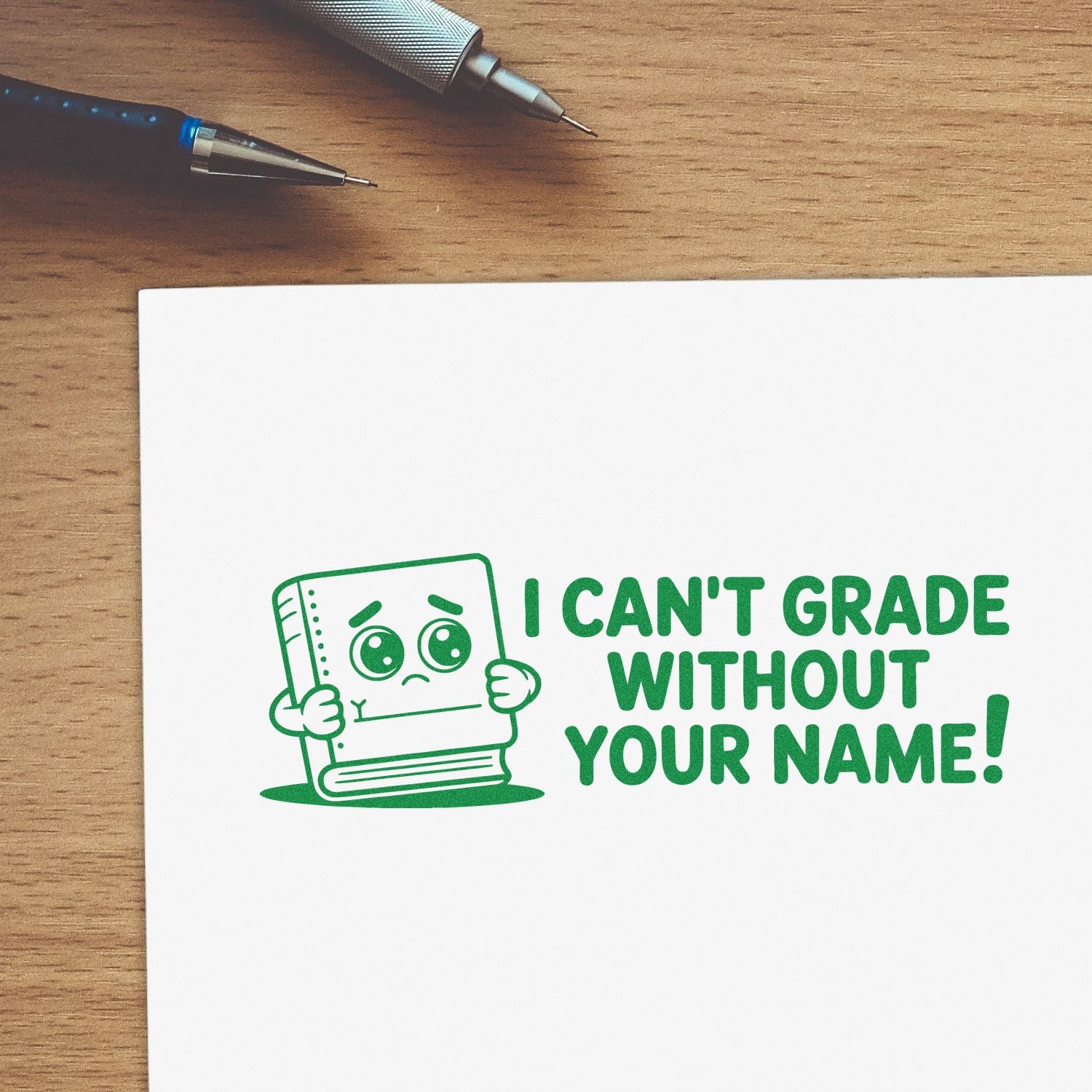 Green custom stamp seal on white paper with cartoon book and I CANT GRADE WITHOUT YOUR NAME! beside pens on a wood desk.