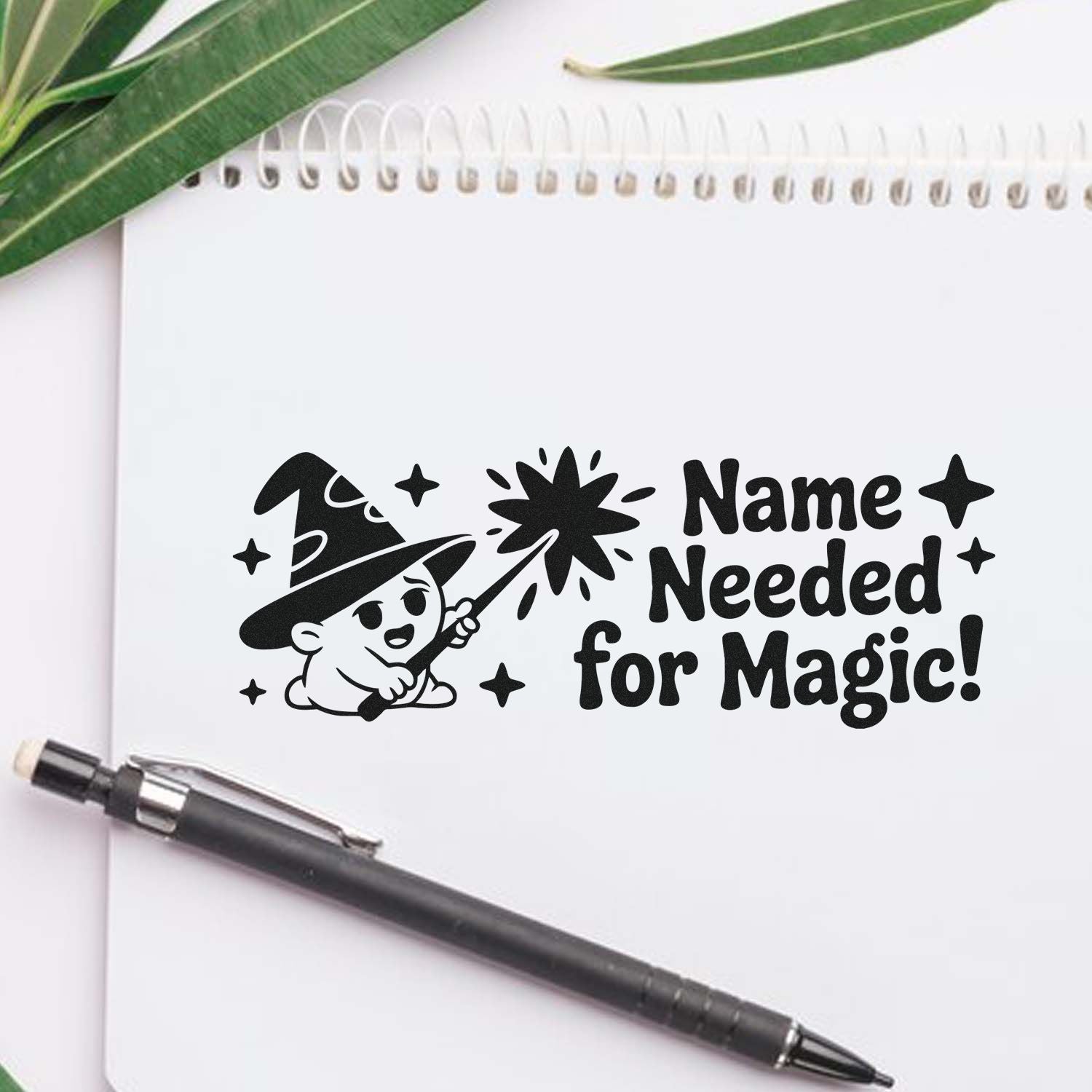 Black custom stamp seal with wizard and sparkles reading Name Needed for Magic! on a white notebook, with pen and green leaves.
