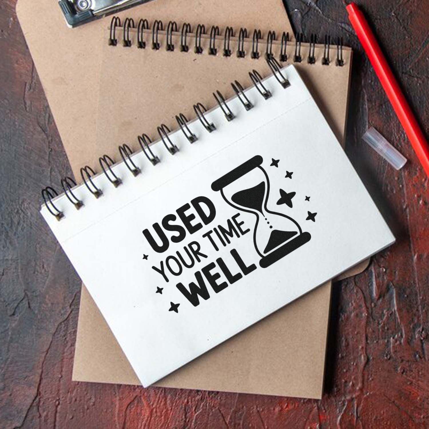 Custom black stamp design on white spiral notebook with hourglass and Used Your Time Well text on kraft paper background.