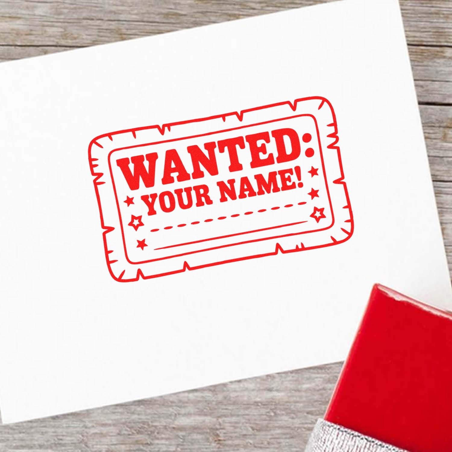 Red wanted-your-name rubber stamp and seal on white paper, with a red stamp pad on a wood surface.
