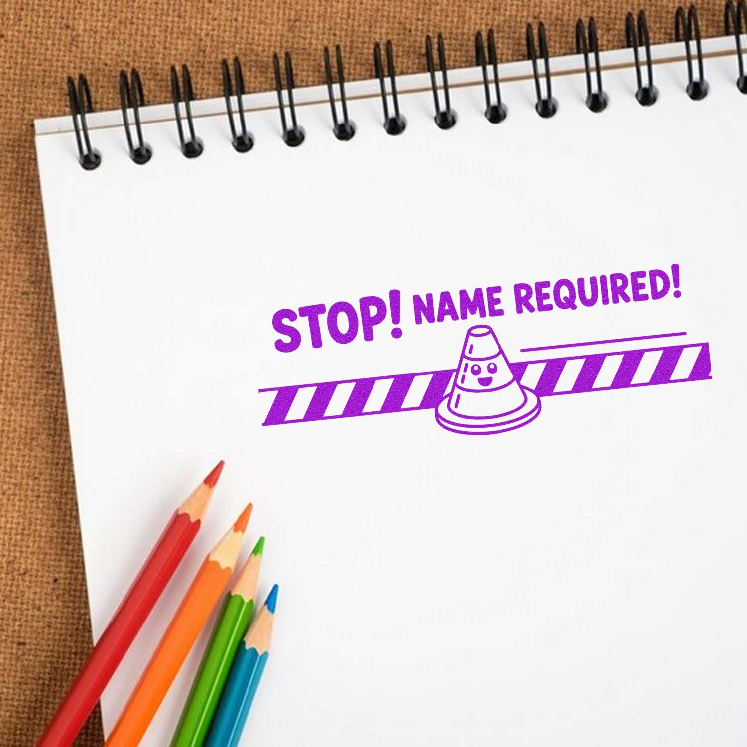 Purple stamp seal reading STOP! NAME REQUIRED! on a white spiral notebook, with colorful pencils and a traffic cone.