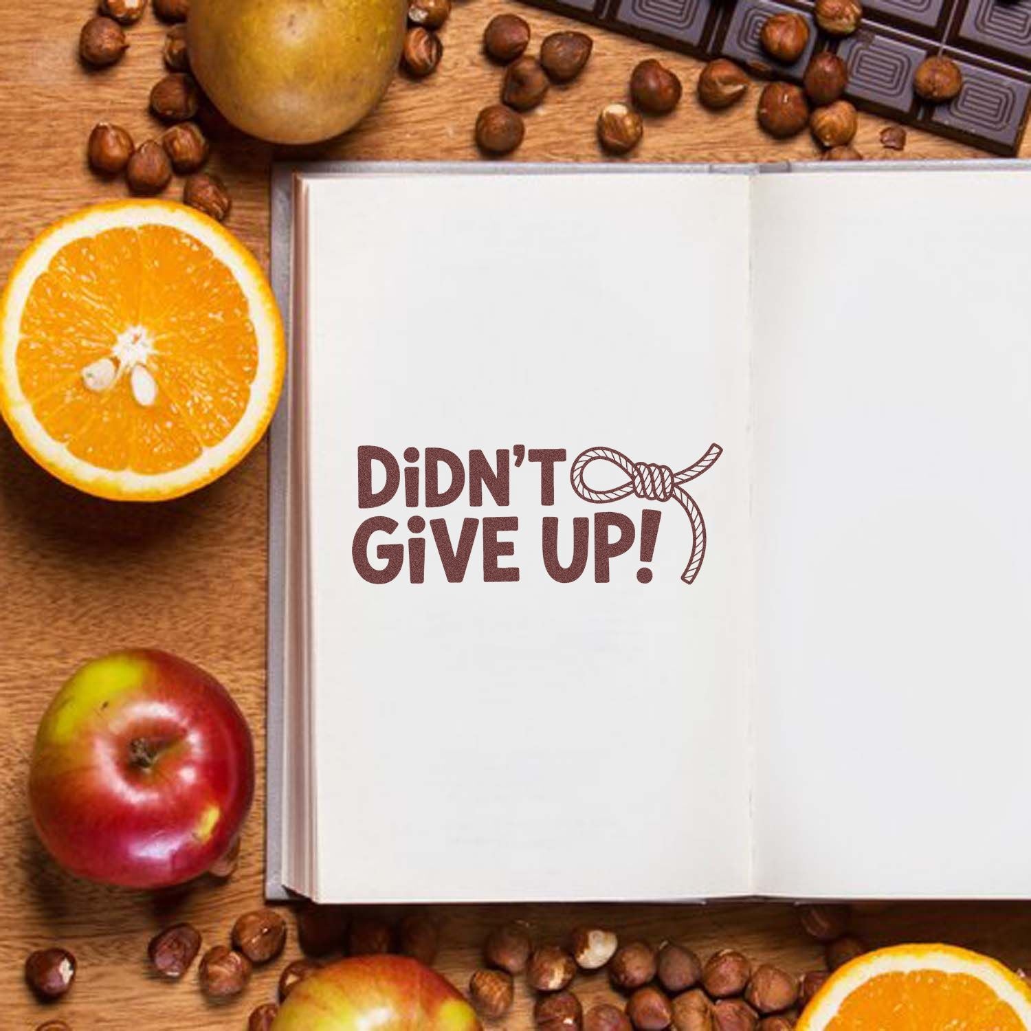 Brown seal stamp reading Didnt Give Up! on an open notebook, with rope knot, oranges, apples, and coffee beans.