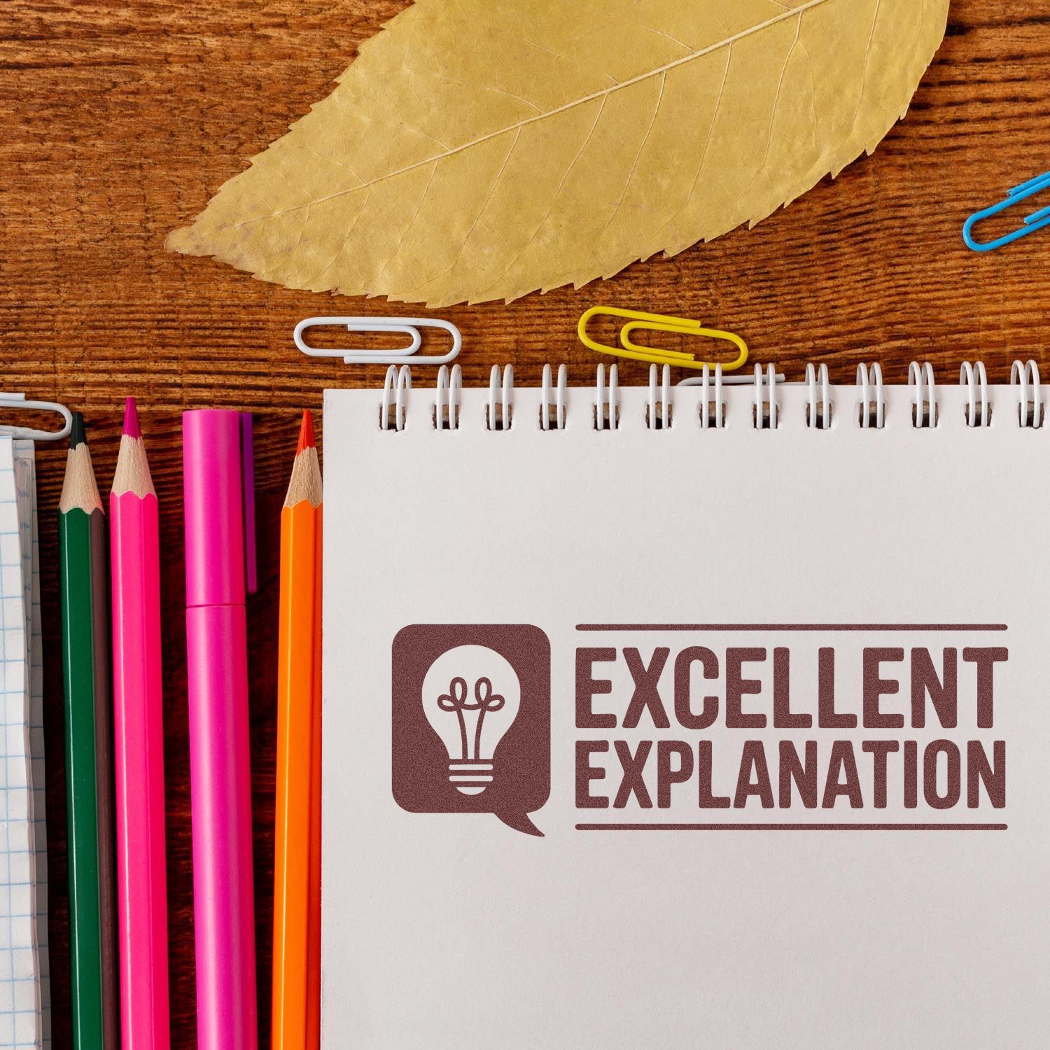 Brown stamp-style seal reading Excellent Explanation on white spiral notebook, with lightbulb icon, colored pencils, and paper clips.