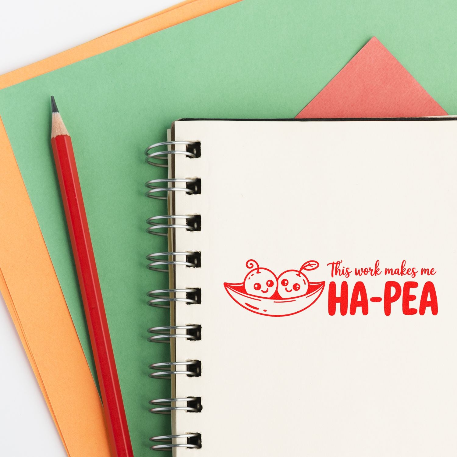 Red stamp seal on spiral notebook reading This work makes me HA-PEA, with pea pod characters, beside a red pencil on green and orange paper.