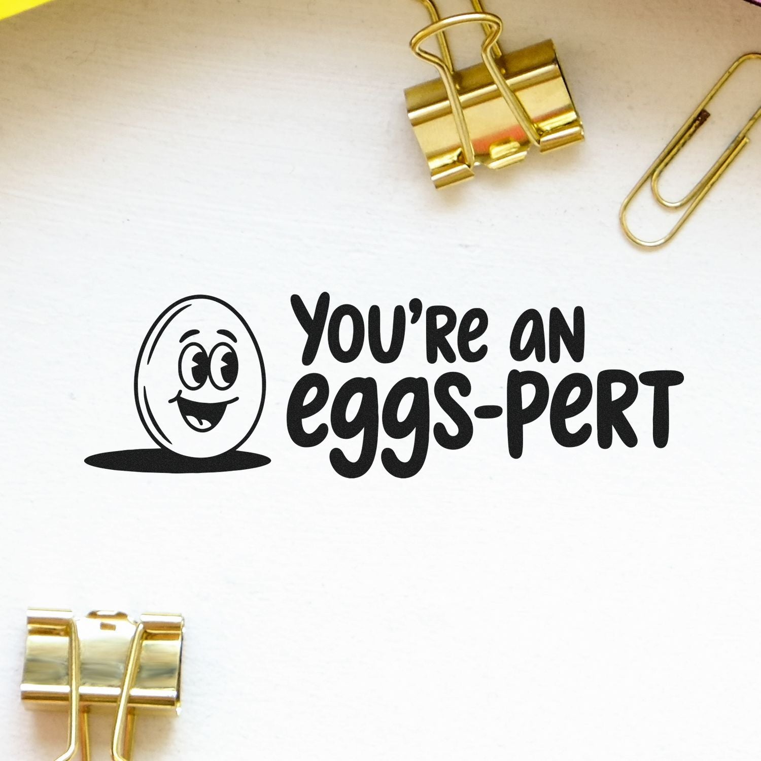 Black stamp-and-seal imprint reading Youre an eggs-pert with smiling egg icon on white paper, framed by gold binder clips.