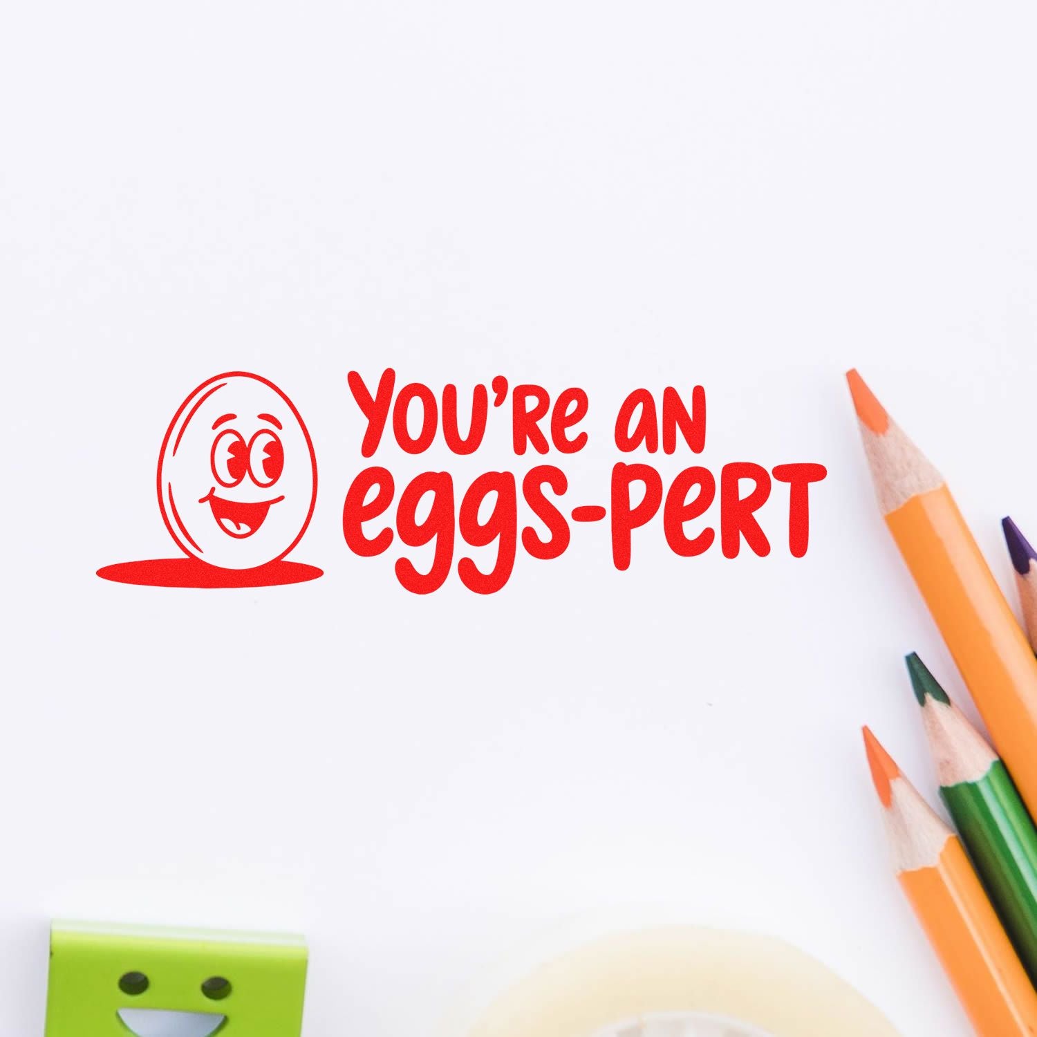 Red stamp seal with smiling egg face and Youre an eggs-pert text on white paper, framed by colored pencils.