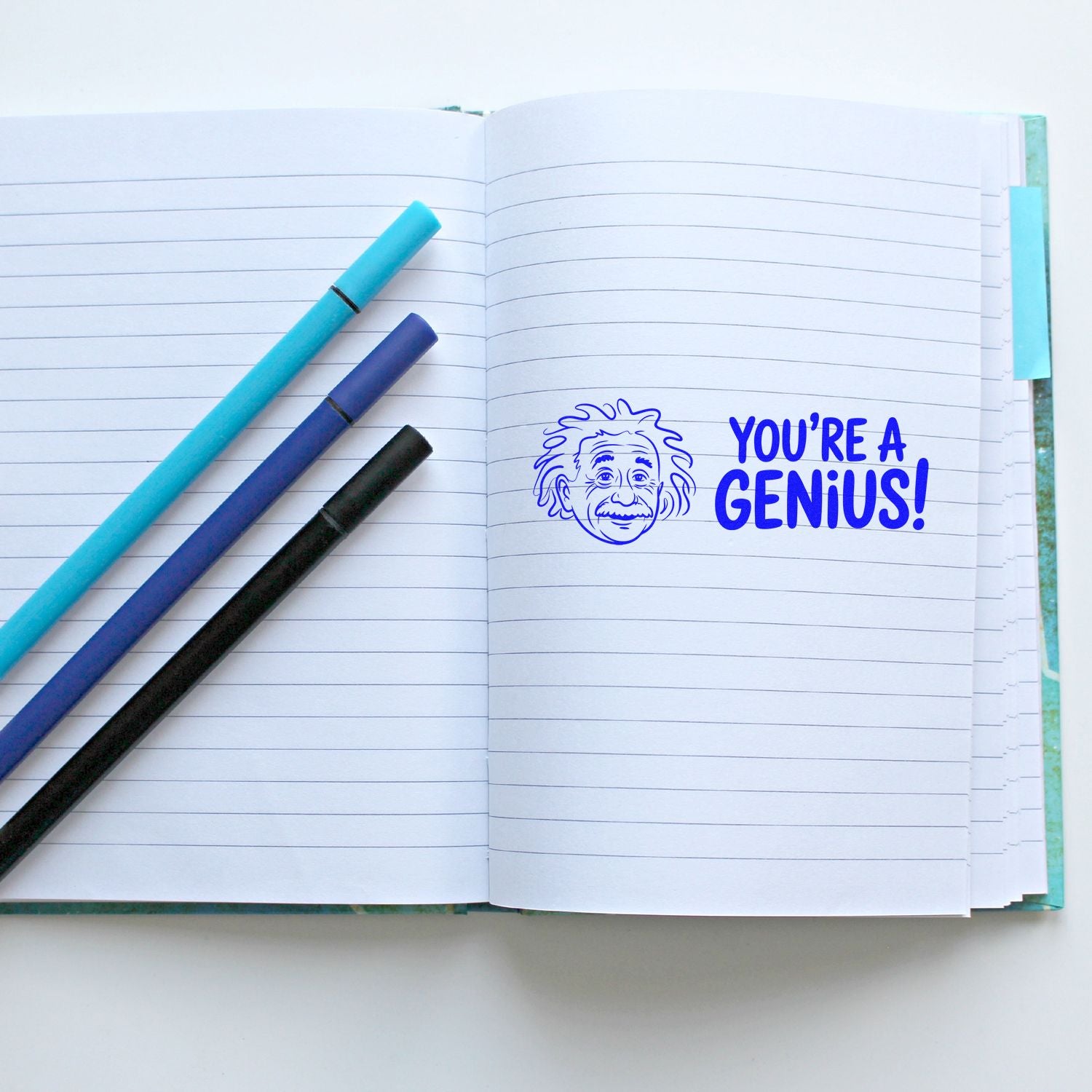 Blue YOURE A GENIUS! stamp and seal with Einstein sketch on lined notebook, beside turquoise, navy, and black pens.