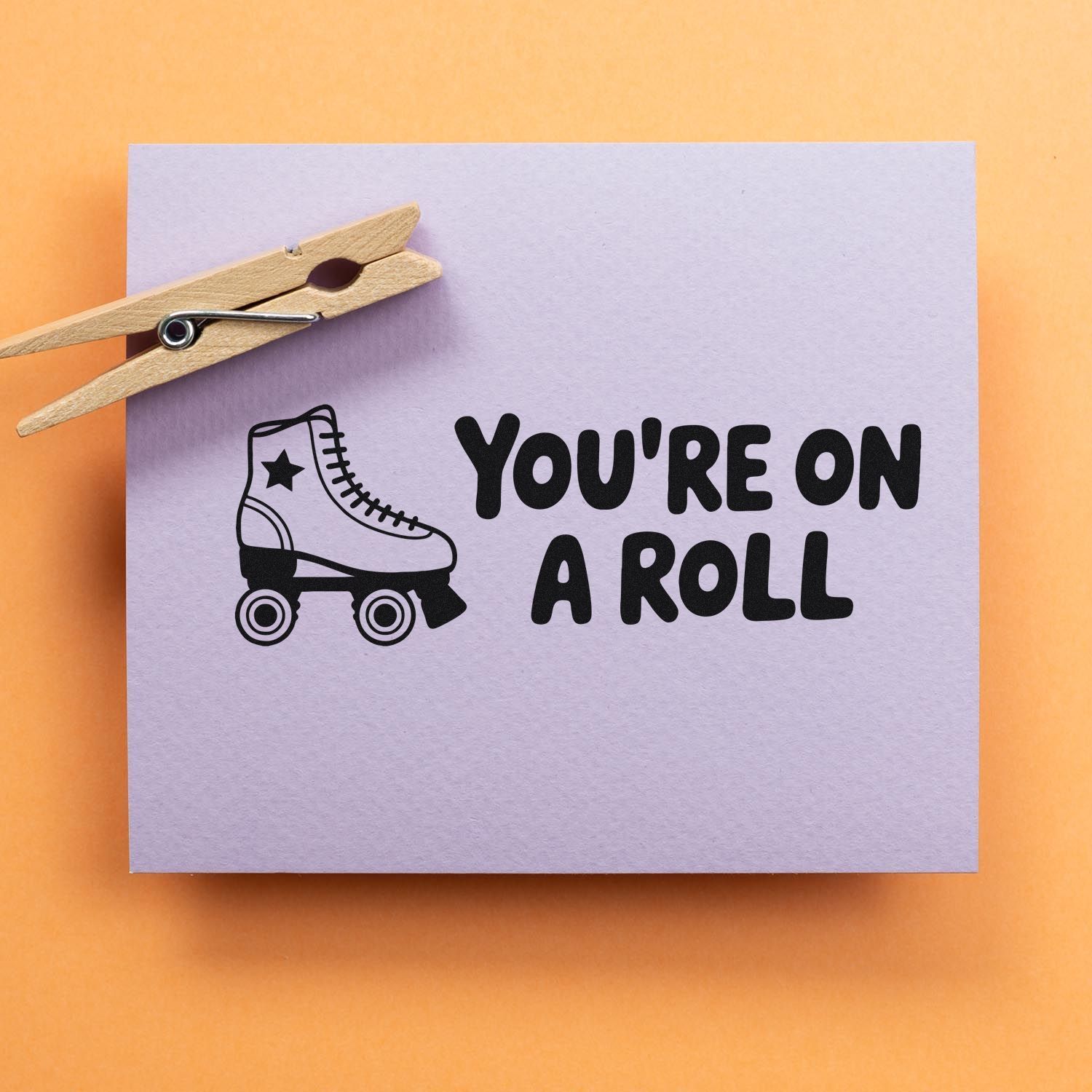 Wooden clothespin holding lavender paper with black roller skate stamp and Youre on a roll text on orange background