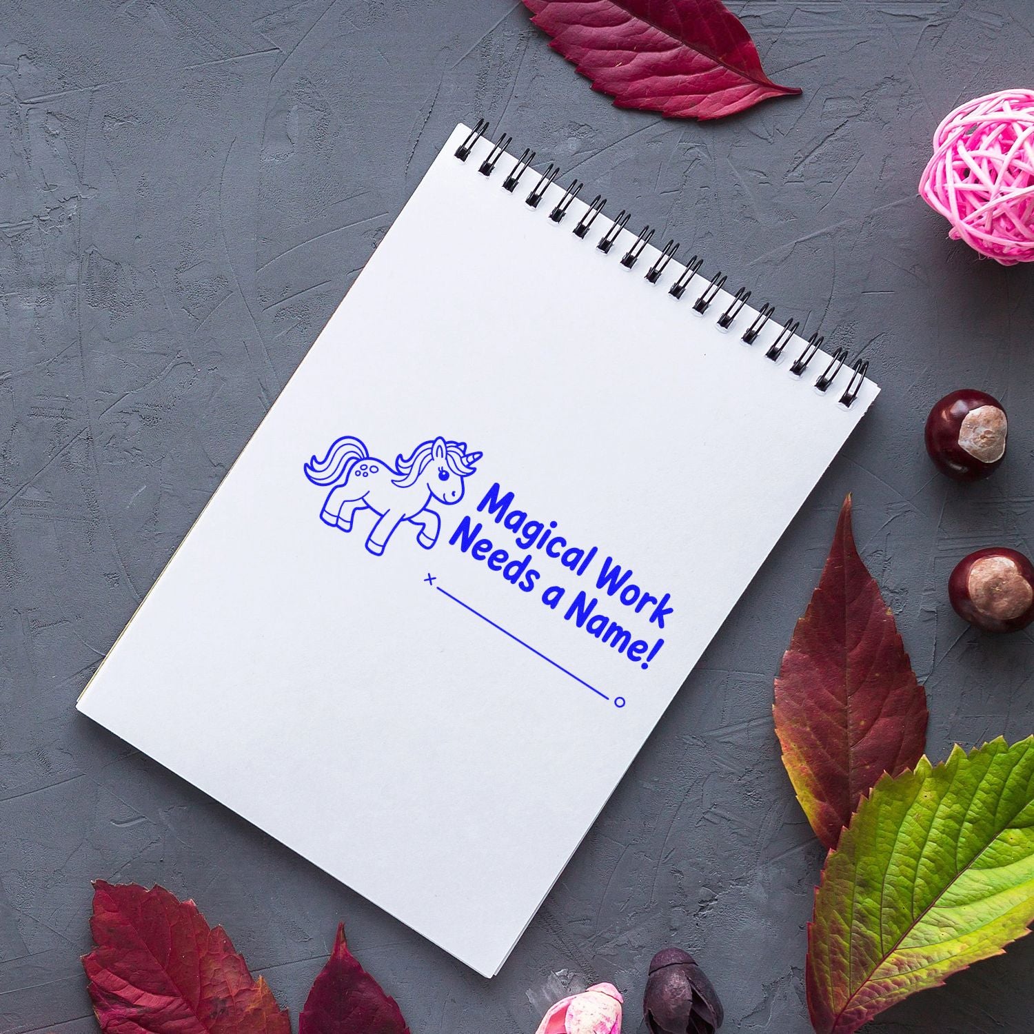 Blue custom stamp on white spiral notebook with unicorn and Magical Work Needs a Name! text, on gray background with autumn leaves.