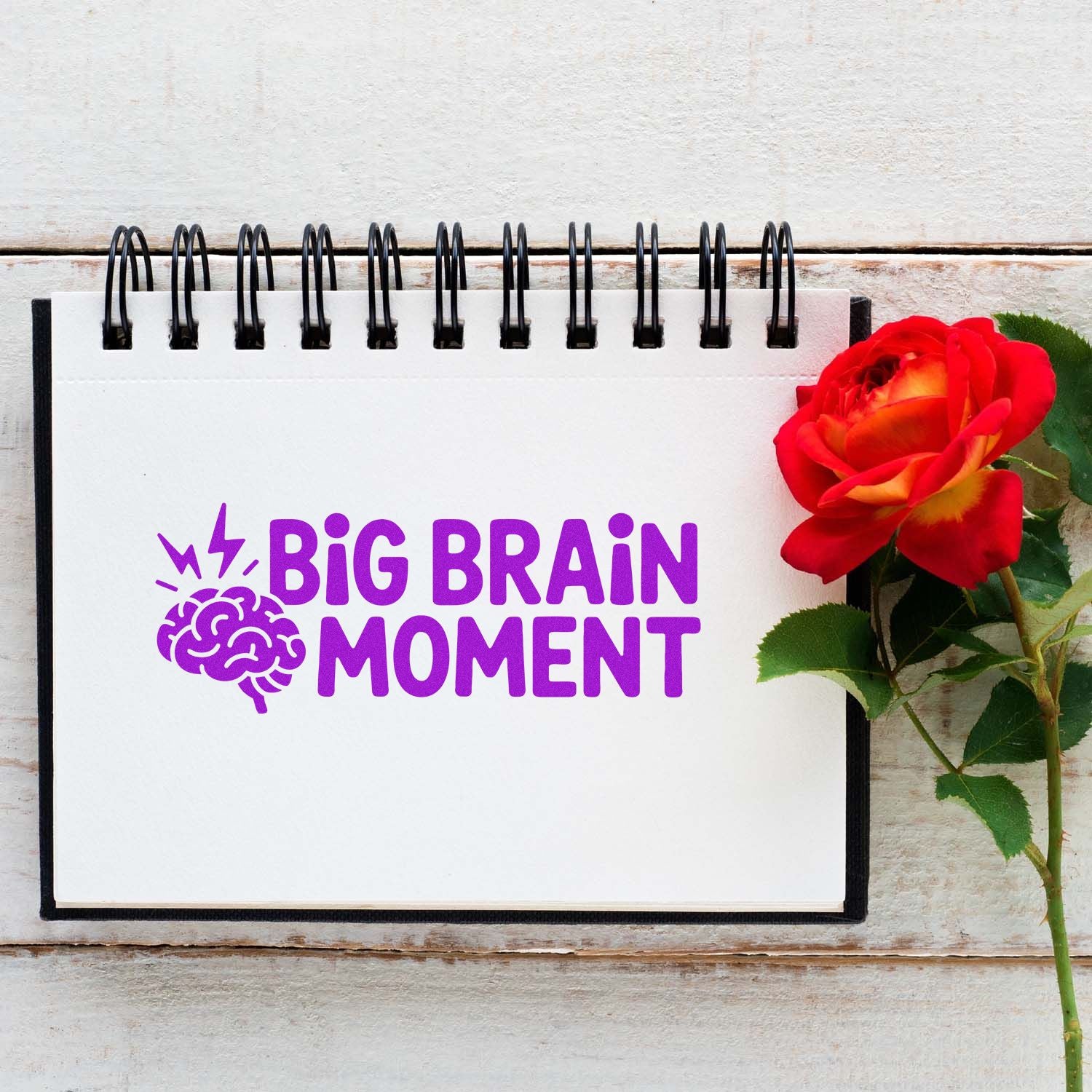 Purple custom stamp and seal design reading Big Brain Moment on a white spiral notebook beside a red rose.