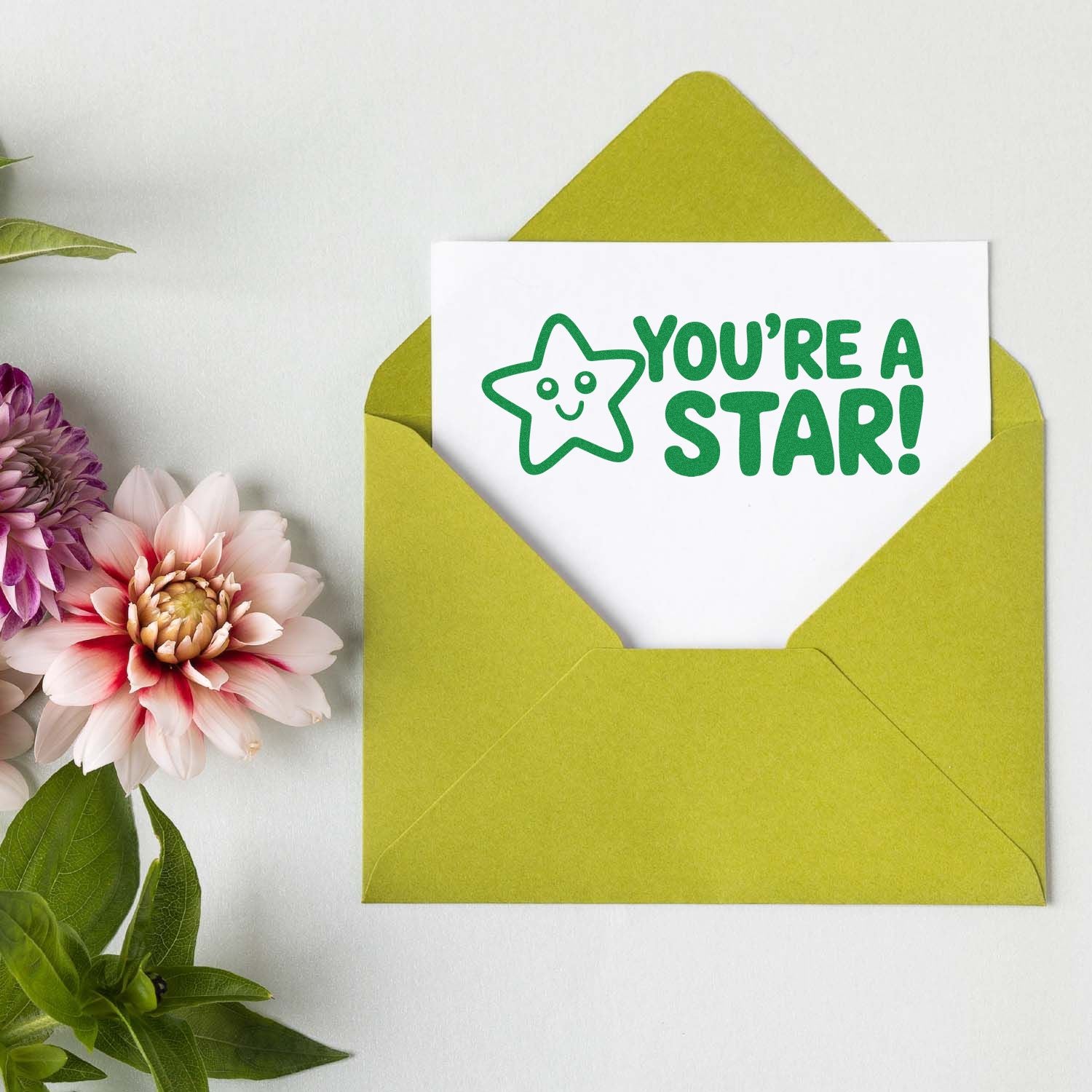 Green envelope with a white card featuring a Youre a Star! stamp seal and smiling star, beside pink and purple flowers.