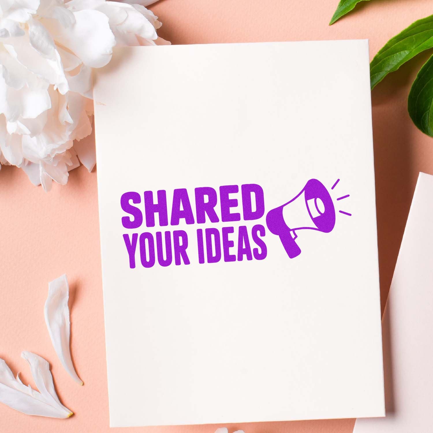 Purple rubber stamp seal reading Shared Your Ideas with megaphone on white paper, on a peach background with white petals.