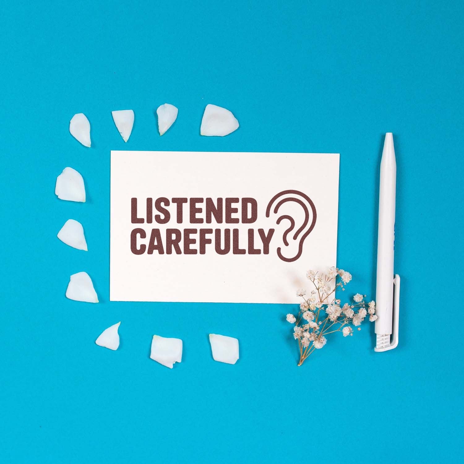 White stamp card reading LISTENED CAREFULLY with ear icon on blue background, surrounded by white seal pieces, pen, and dried flowers.