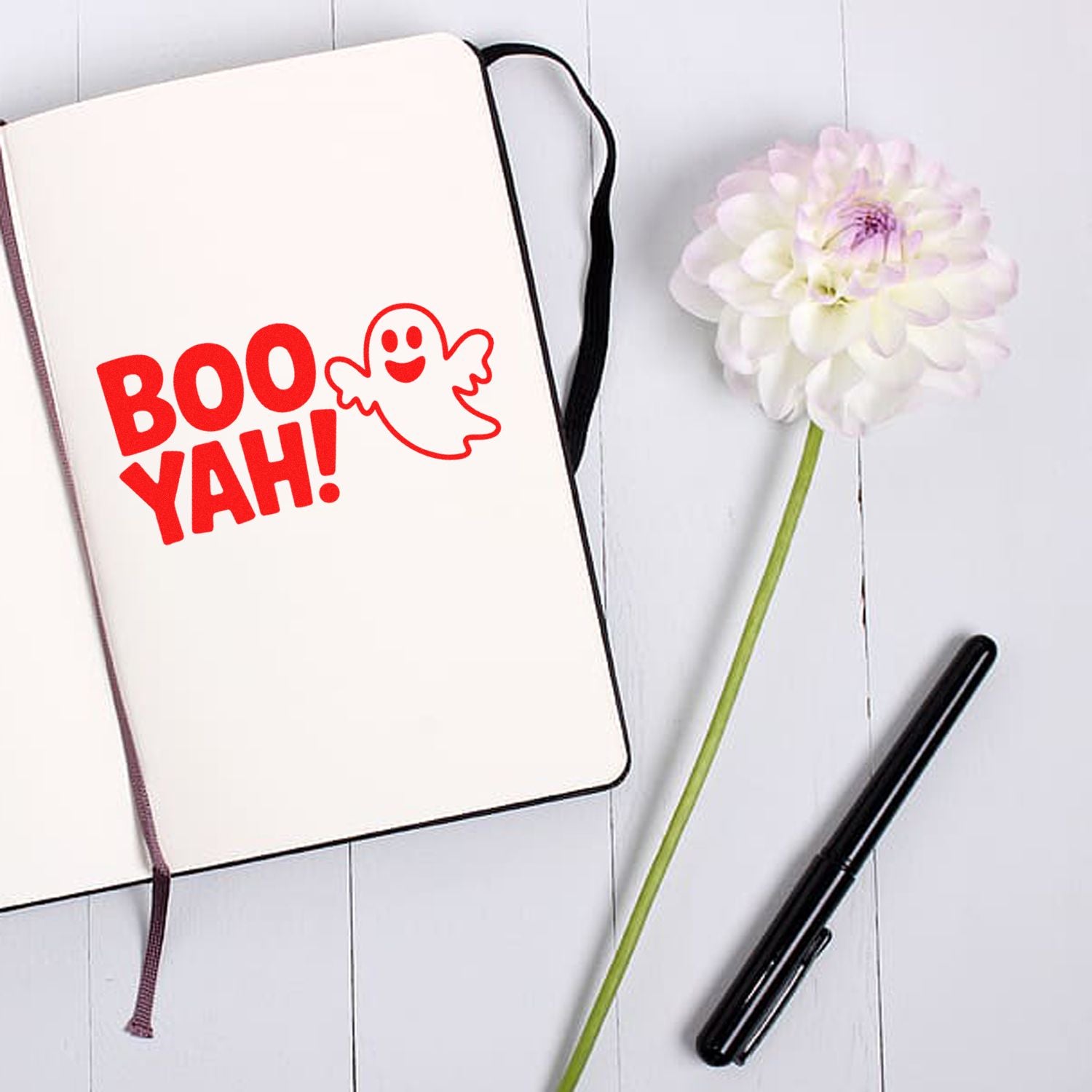 Red BOO YAH! stamp and seal design in a cream notebook, with ghost icon, black pen, and white flower on a white desk.