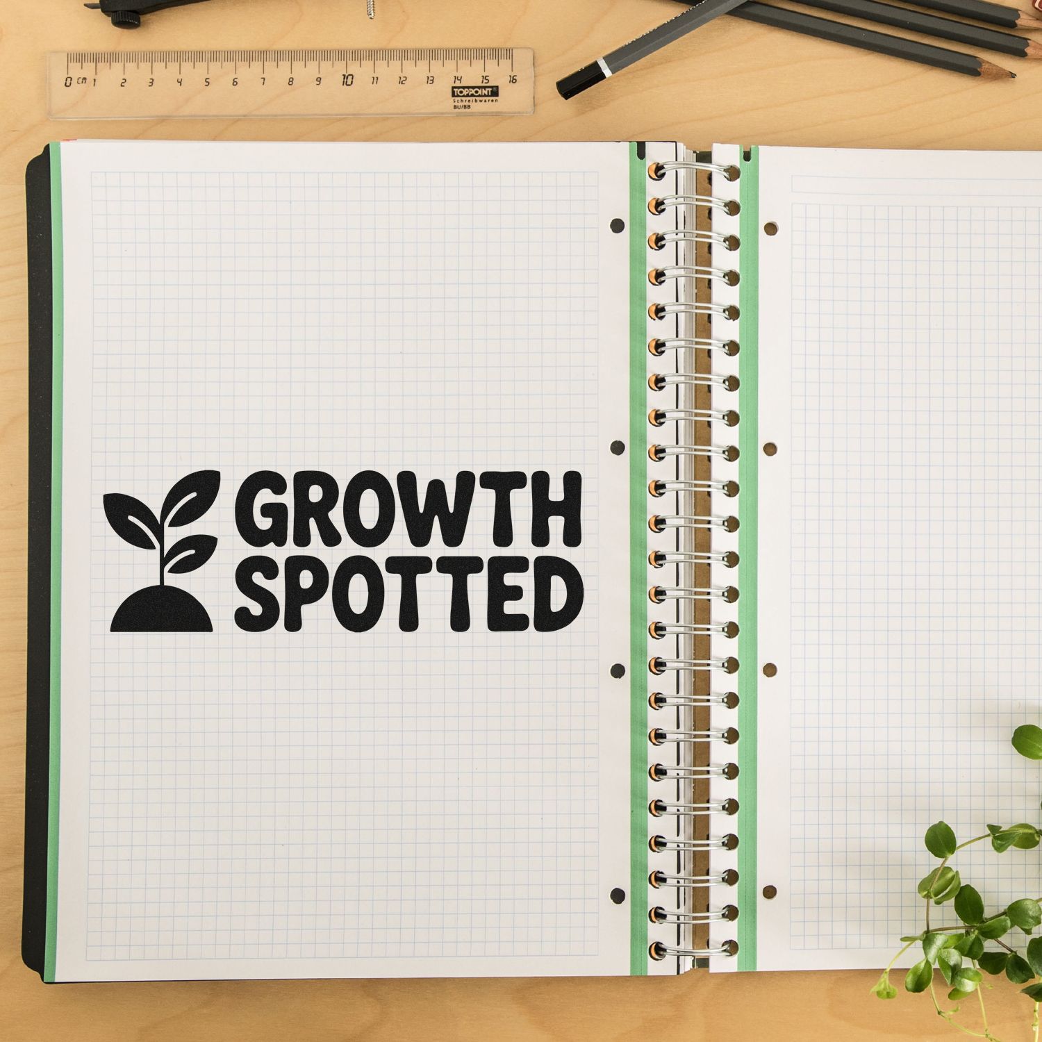 Black rubber growth stamp and seal on open graph notebook with green accents, ruler, pencils, and wooden desk.