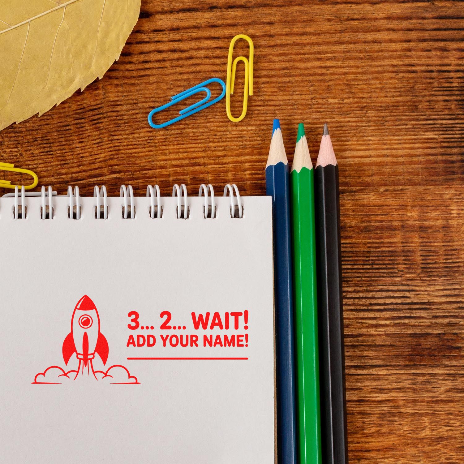 Red custom stamp on notebook reading 3...2... WAIT! ADD YOUR NAME! with blue, green, and black pencils on wood desk.