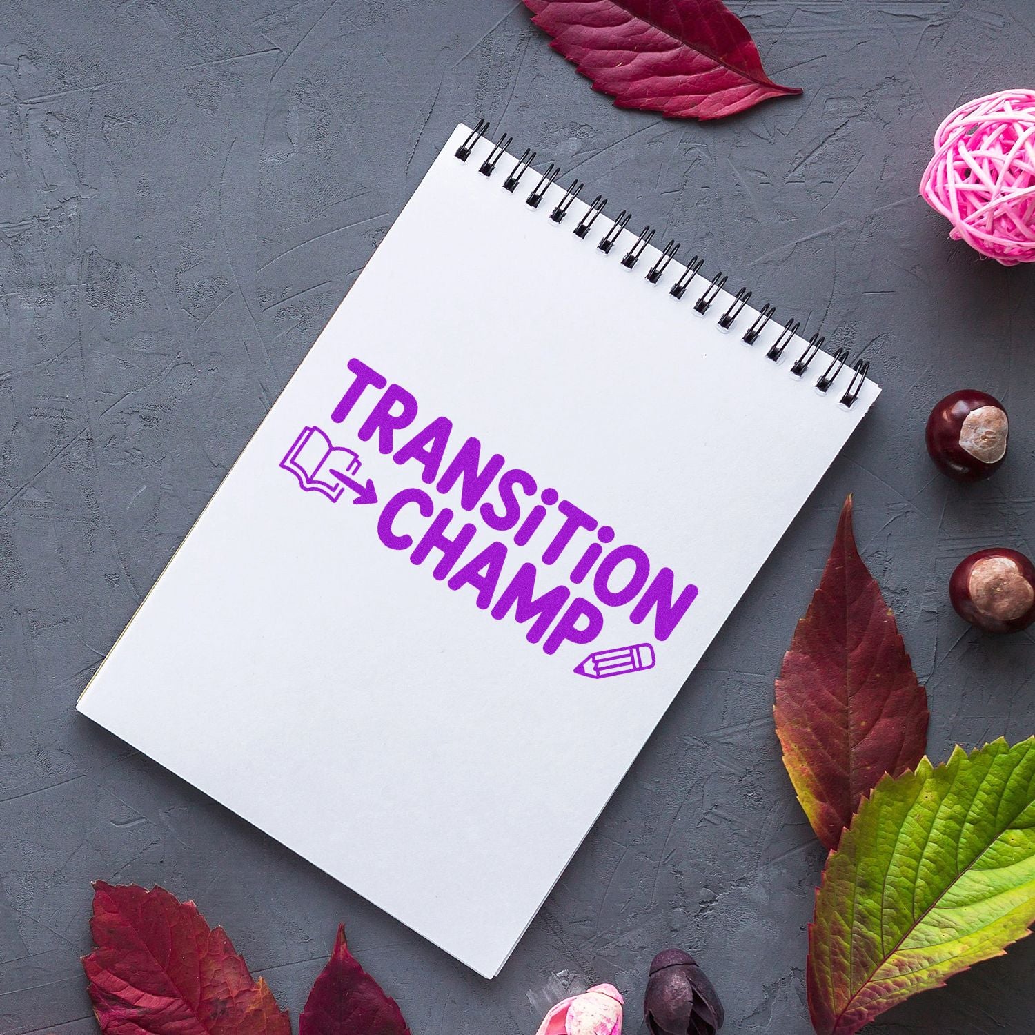 White spiral notebook with purple TRANSITION CHAMP stamp-and-seal design, on gray background with autumn leaves and pink accents.