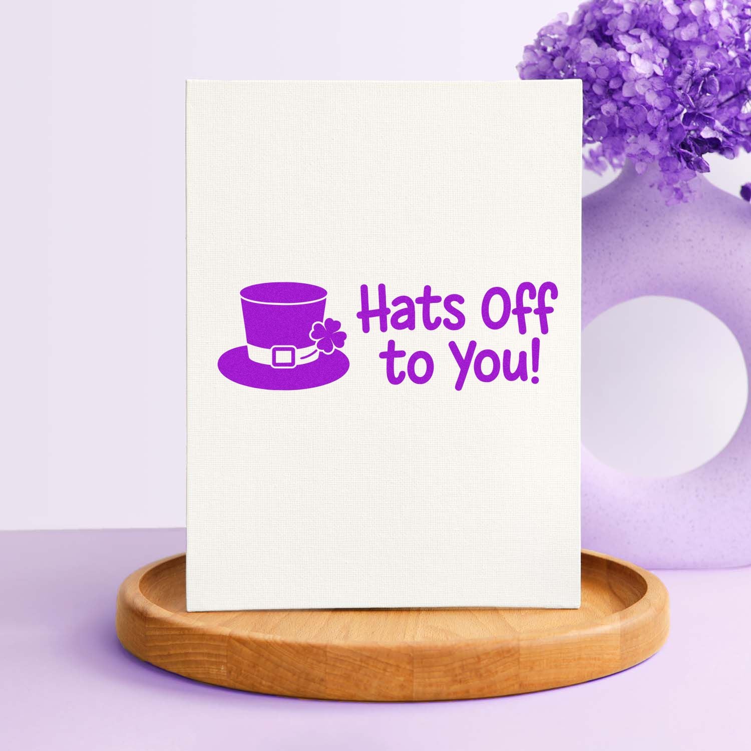 Purple stamp and seal with top hat and clover reading Hats Off to You! on white canvas, atop a wooden base.