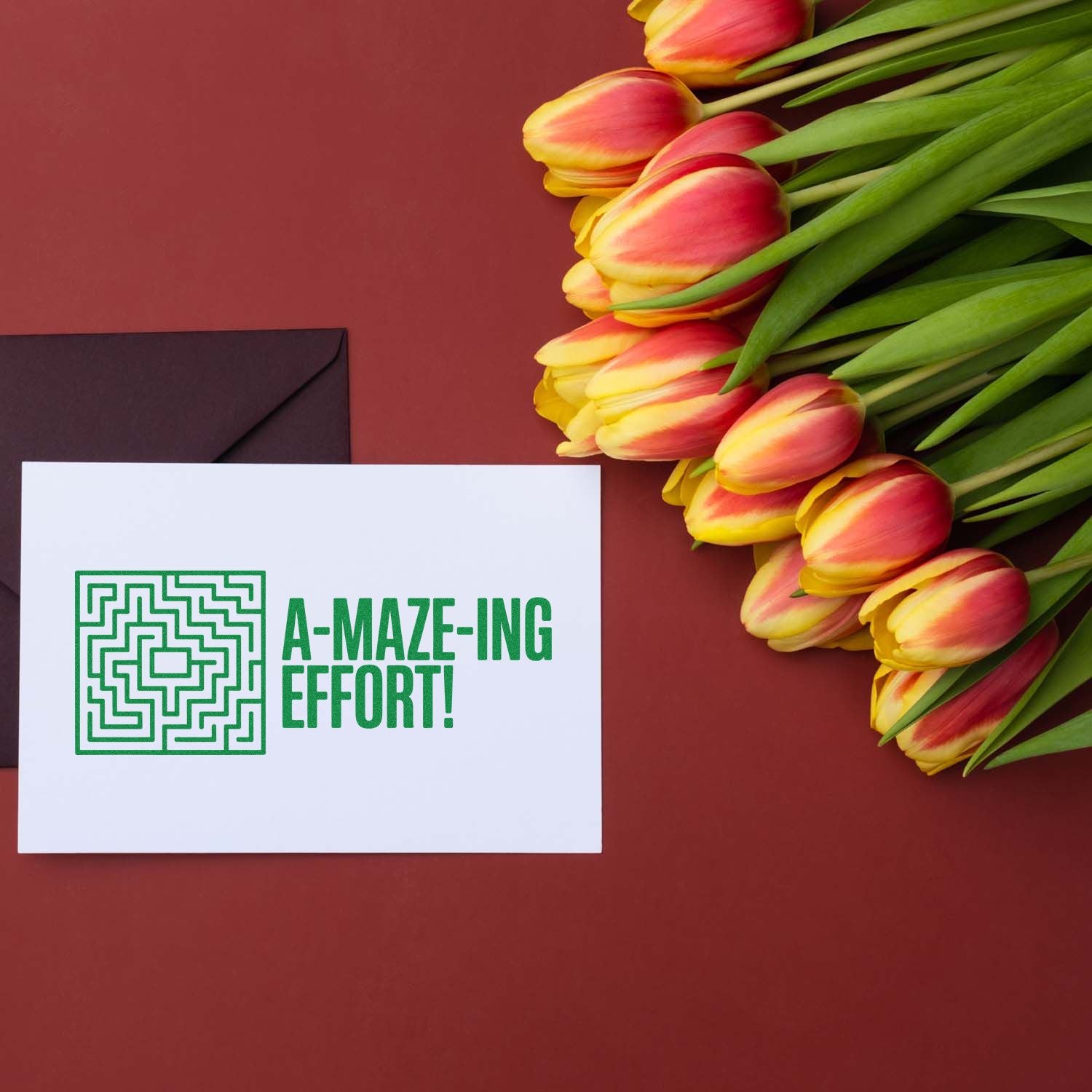 Custom green stamp seal reading A-MAZE-ING EFFORT! with maze icon, beside black envelope and red-yellow tulips on burgundy background.