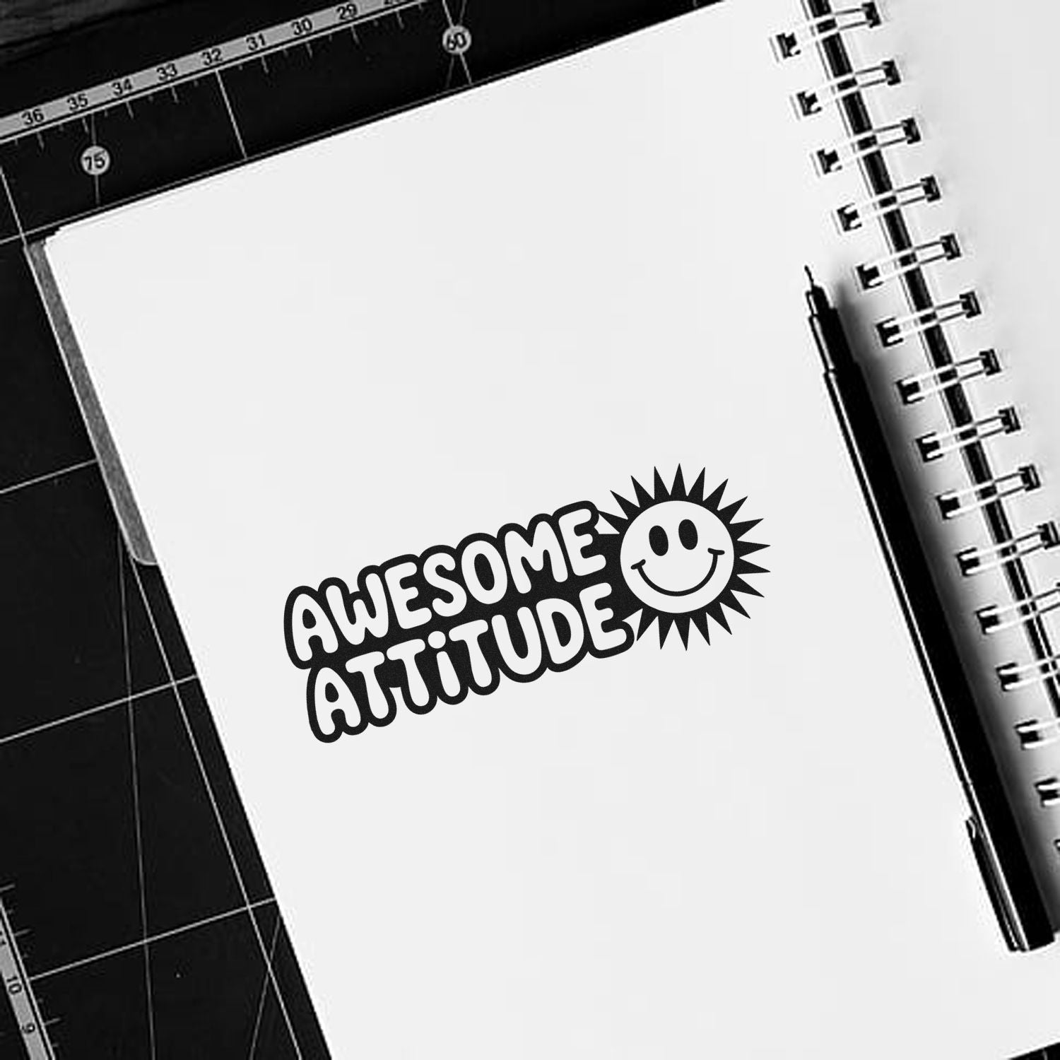 Black Awesome Attitude custom stamp with smiley sun on a white notebook page, beside a spiral binding and pen on a black cutting mat.