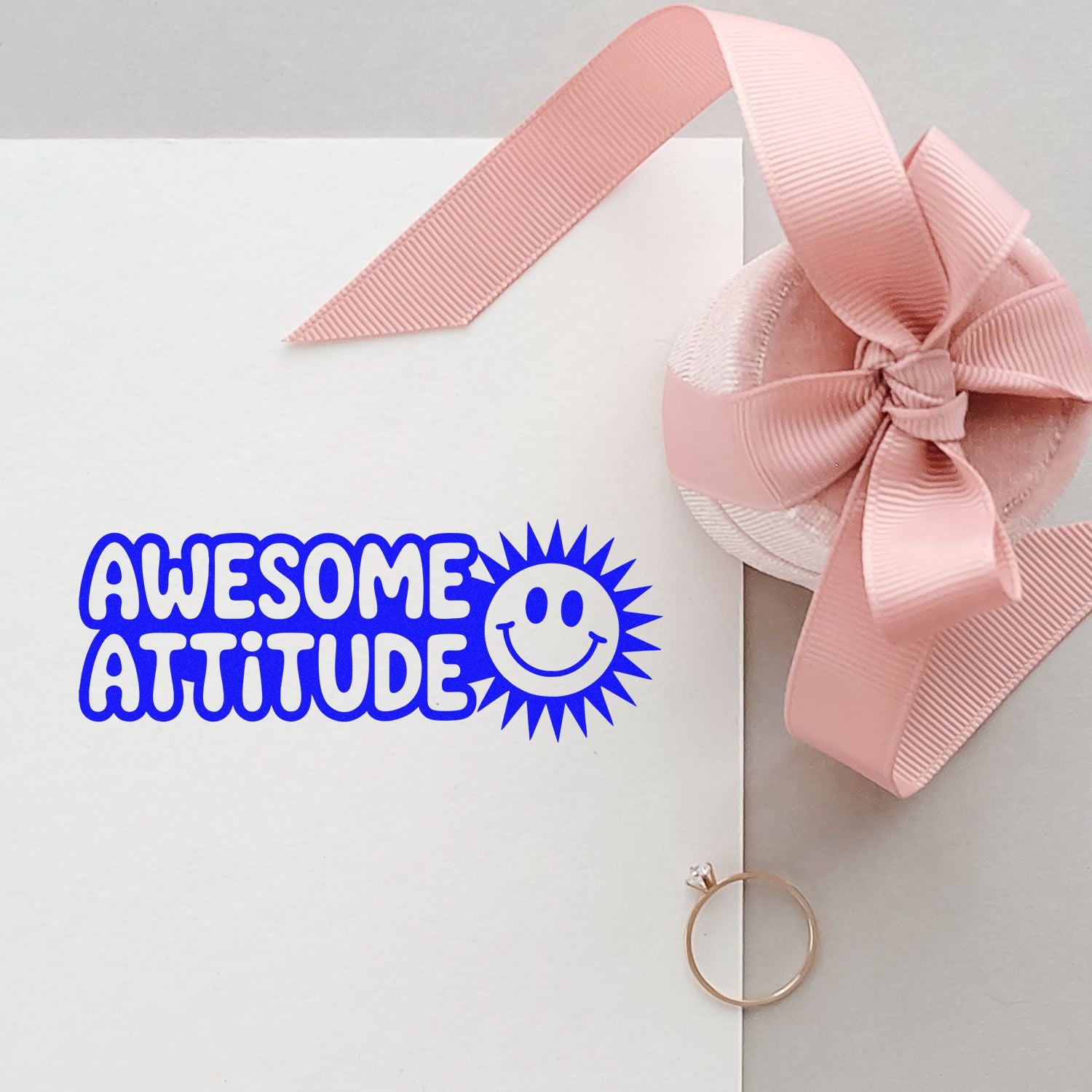 Blue Awesome Attitude stamp with smiley on white card, paired with a pink ribbon seal and gold ring embellishment.