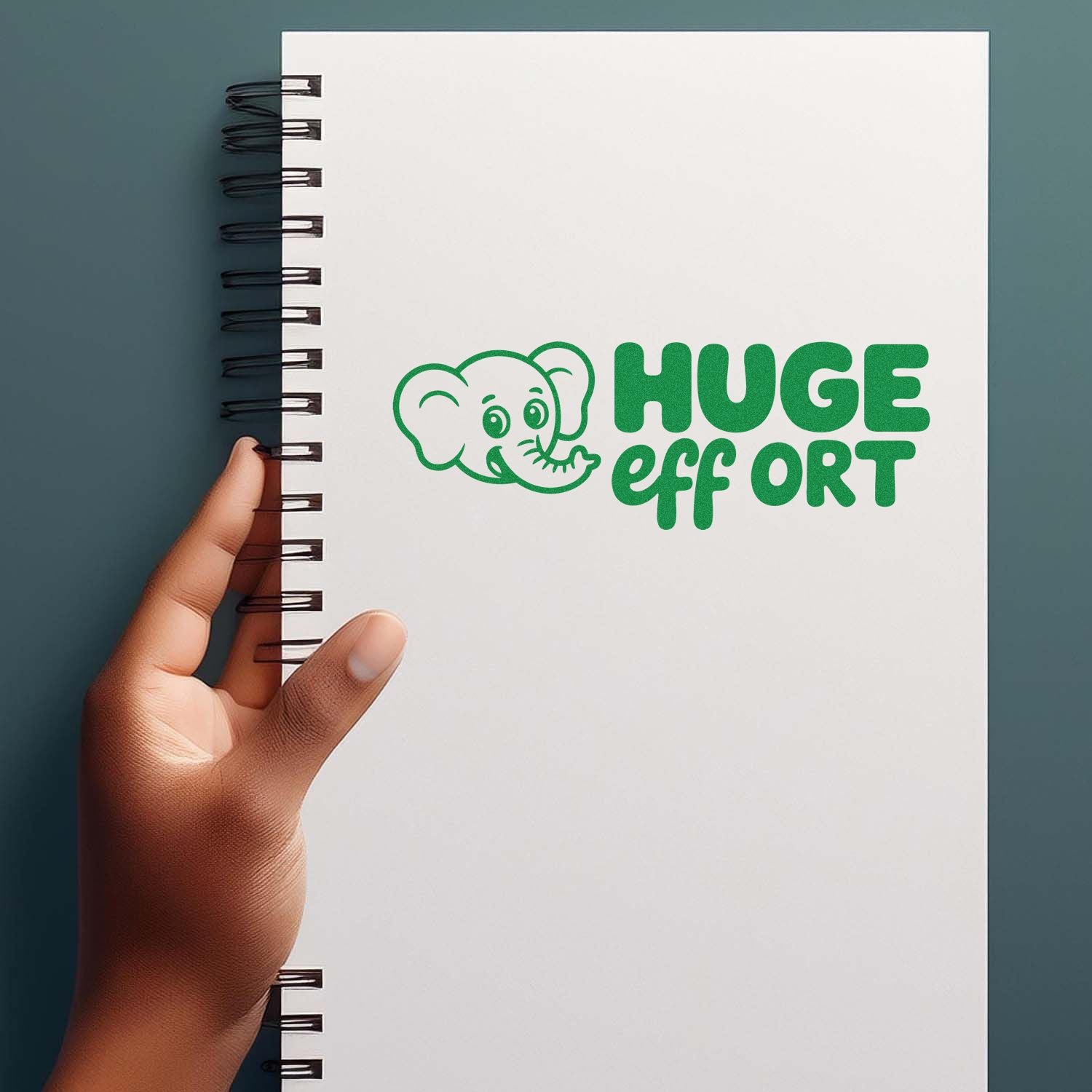White spiral notebook with green elephant HUGE effort stamp seal, held by hand on a teal background.