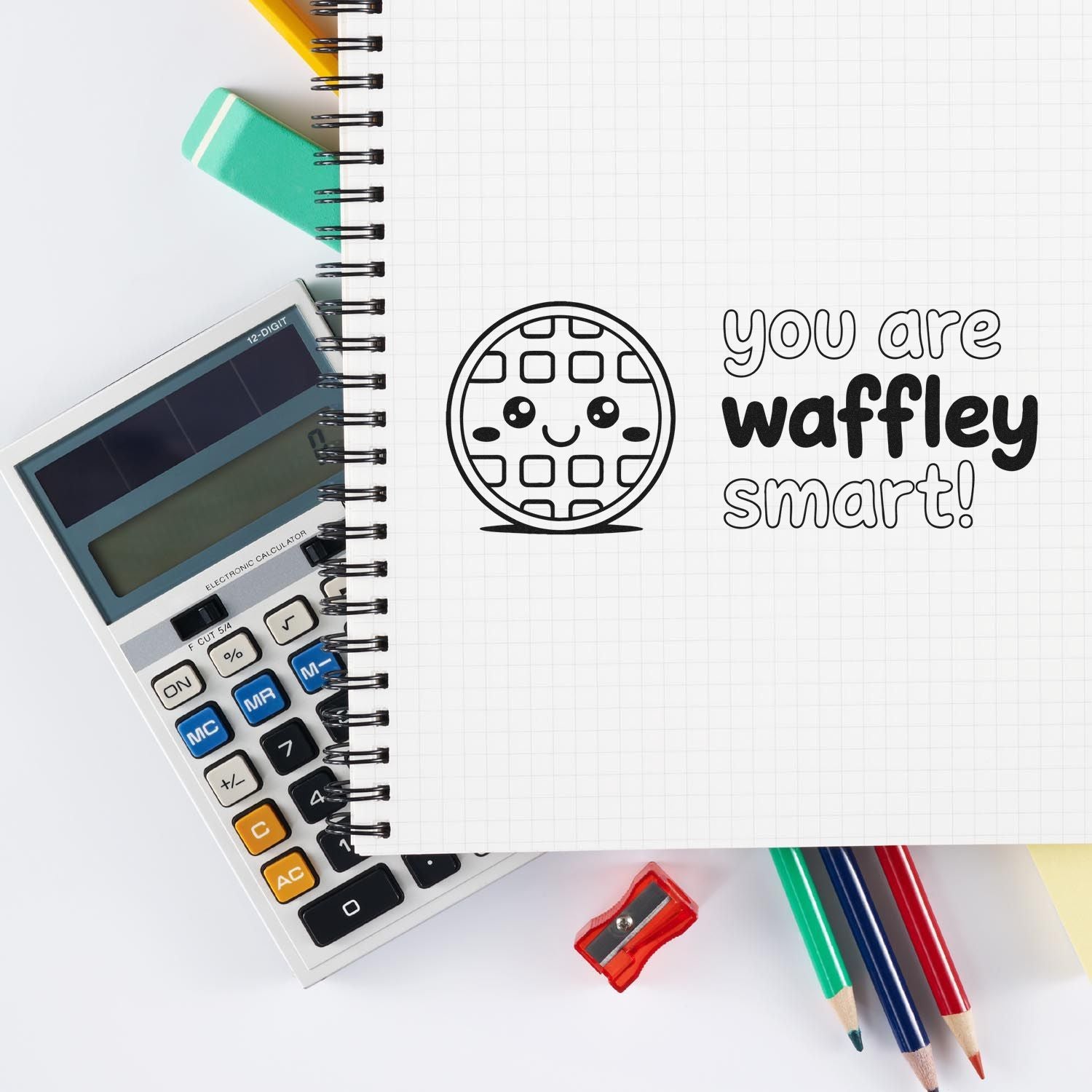 Waffle-shaped rubber stamp with you are waffley smart on a grid notebook, with calculator, pencils, and sharpener.