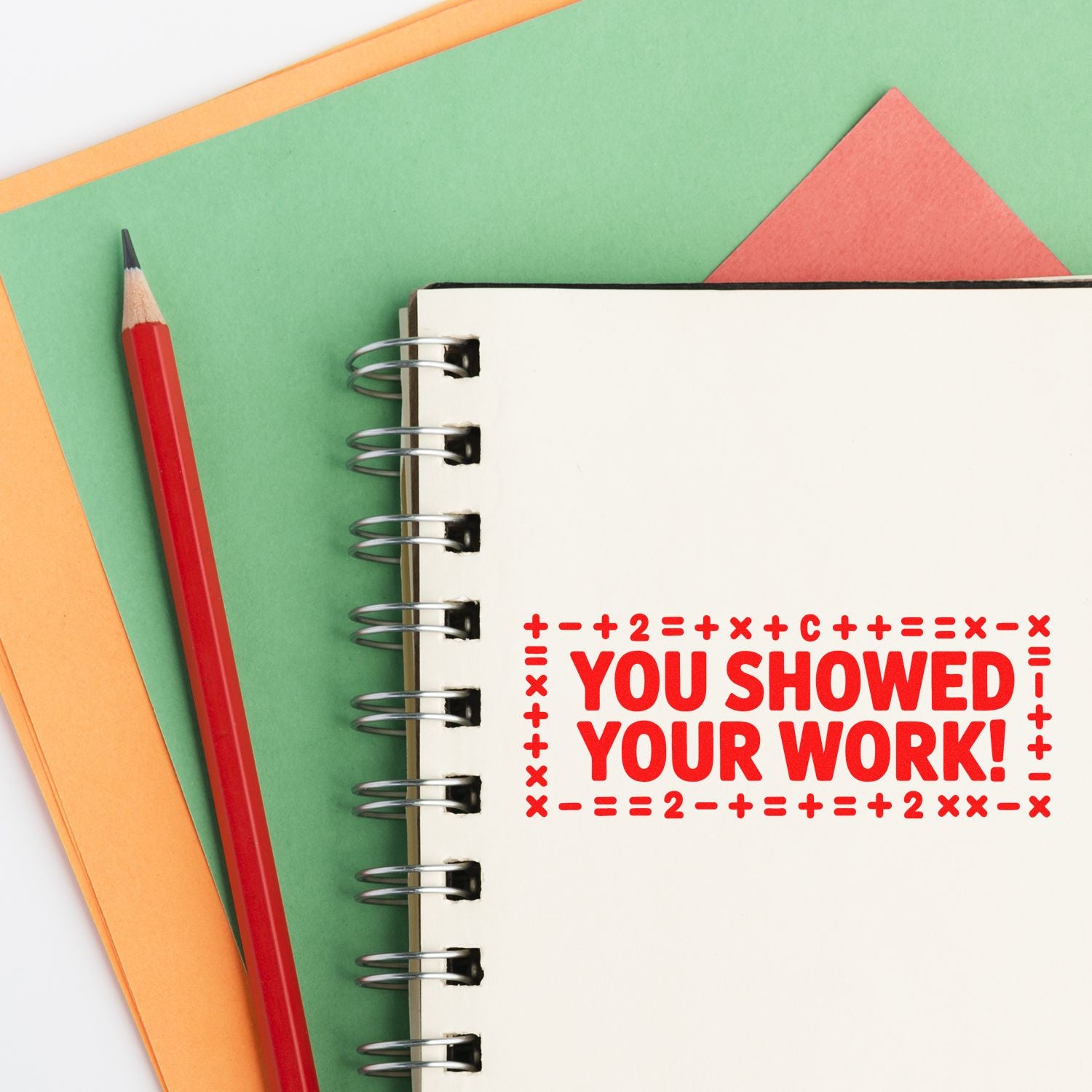 Red rubber stamp seal reading You showed your work! on a spiral notebook, with a red pencil and green/orange paper.