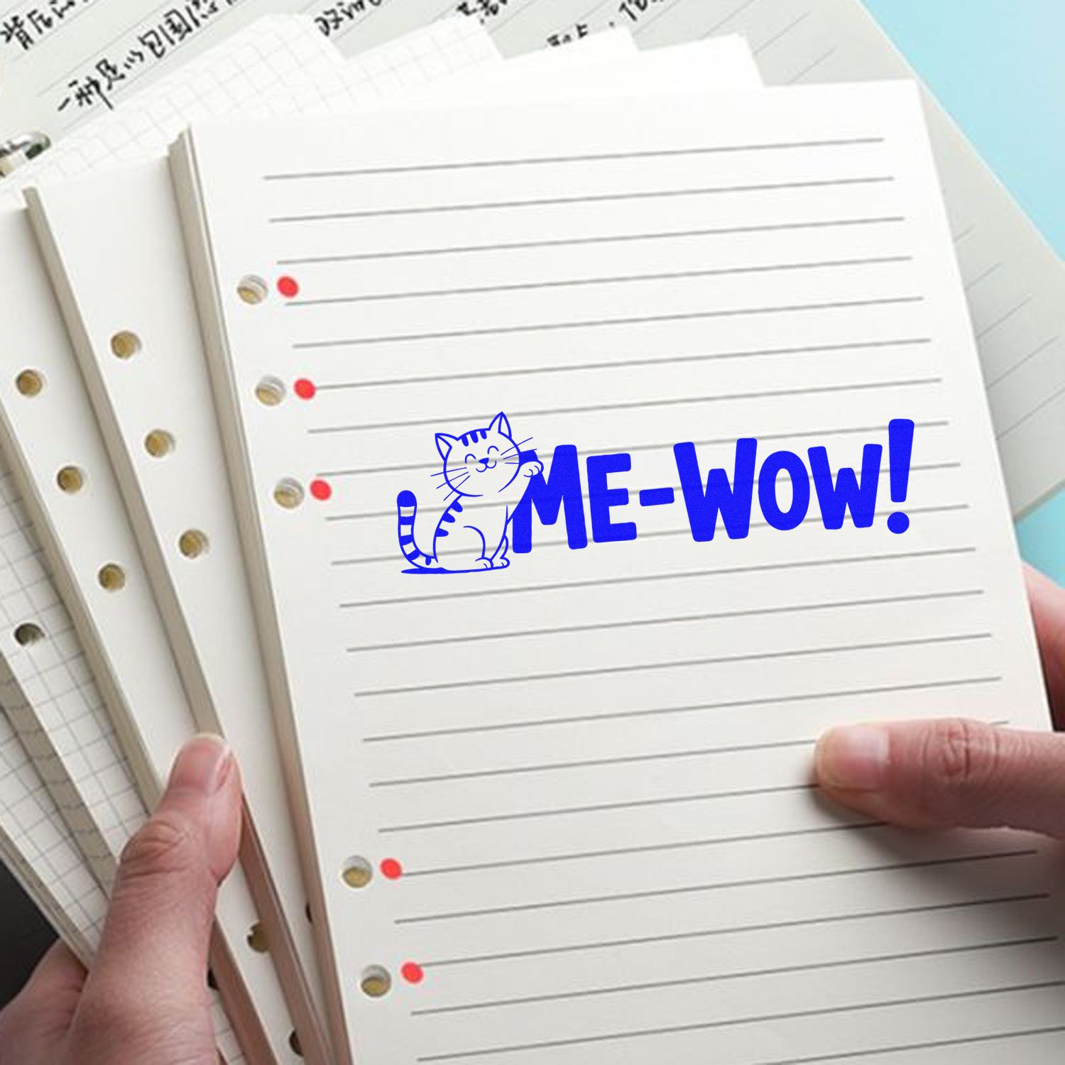 Blue cat ME-WOW! stamp seal on white lined notebook refills, held by hands, with red and gold binder holes.