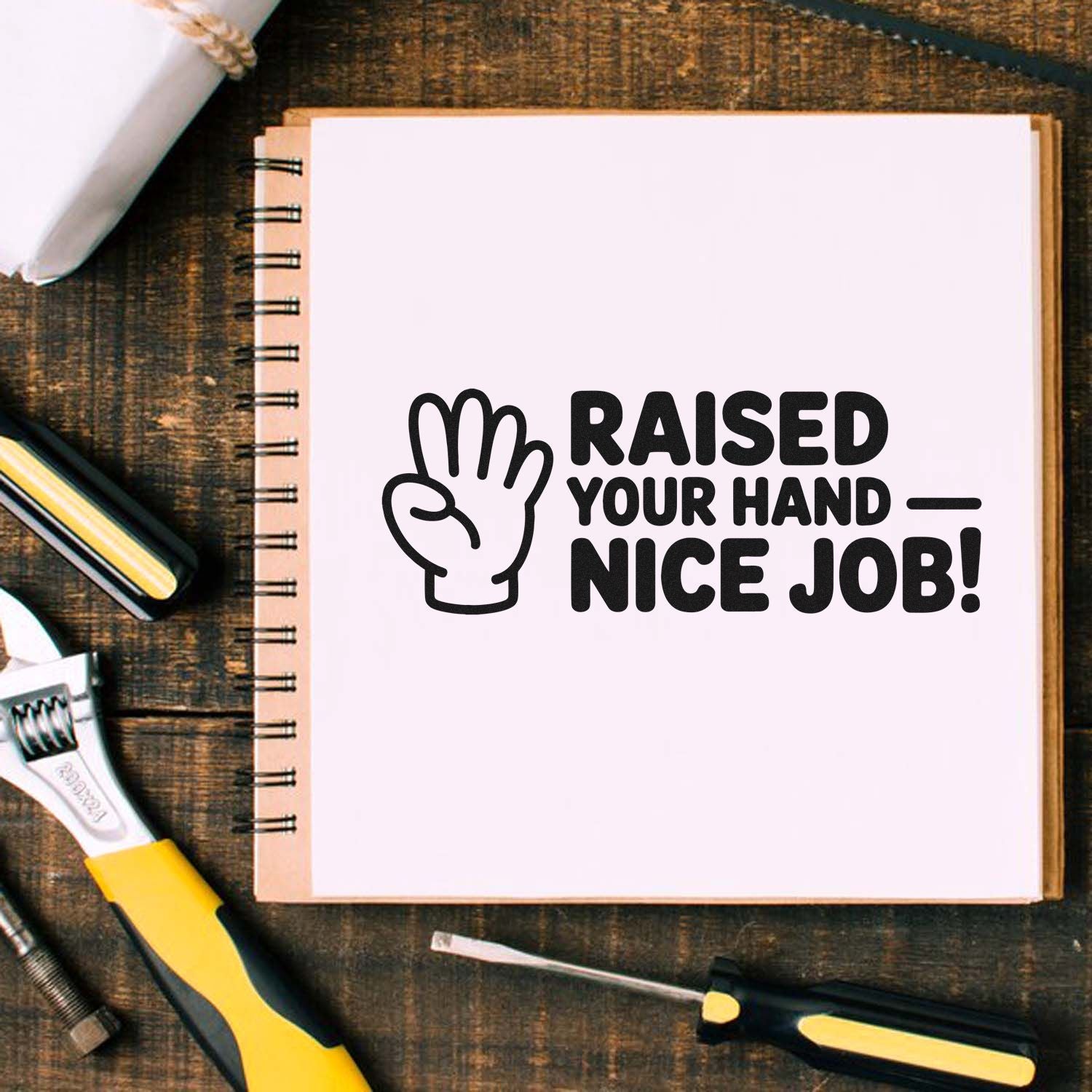 Black hand stamp seal reading Raised Your HandNice Job! on a white spiral notebook with yellow-and-black tools.