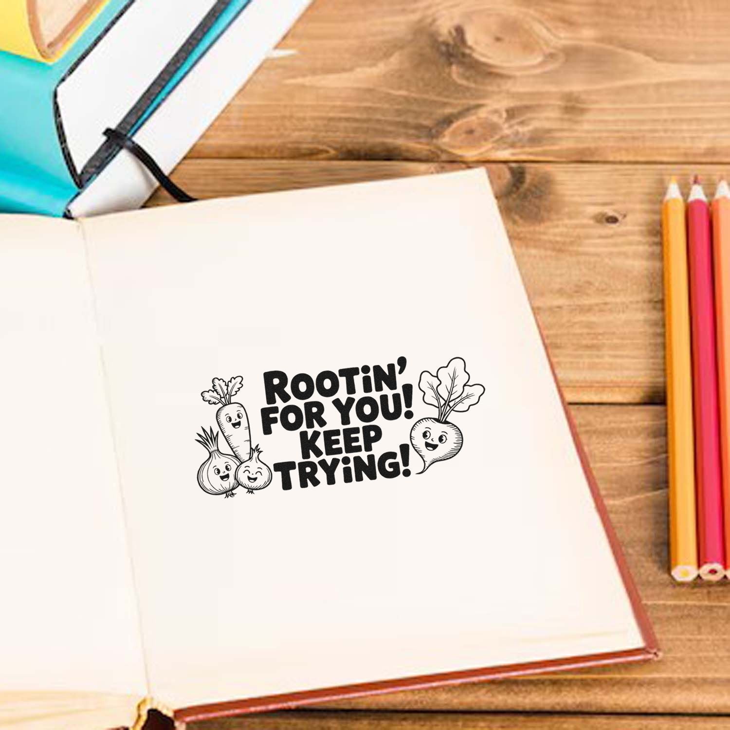 Black stamp seal saying Rootin for you! Keep trying! with cute veggie characters on a white notebook, beside colored pencils.