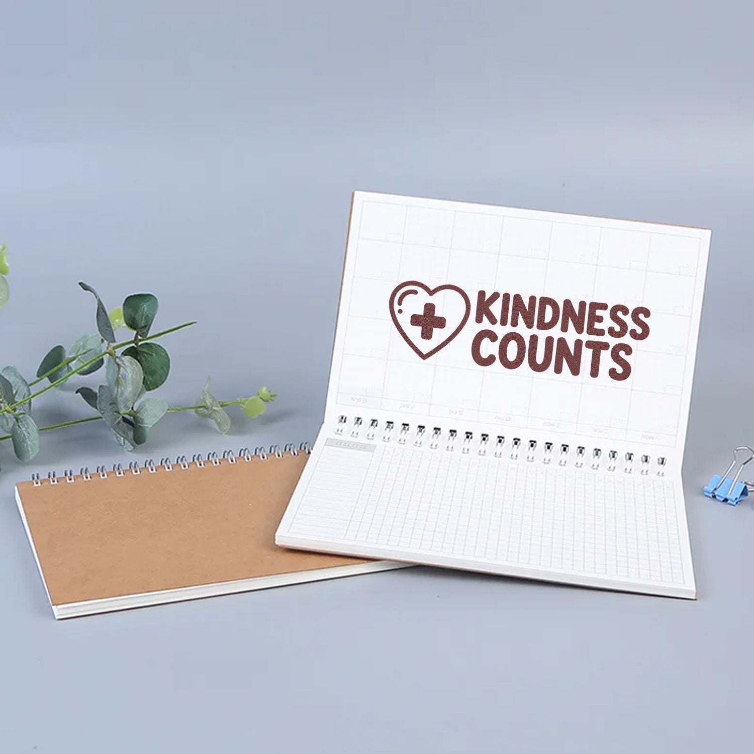 White stamp and seal notebook with brown Kindness Counts heart logo, beside a kraft spiral notebook, blue clip and green leaves.