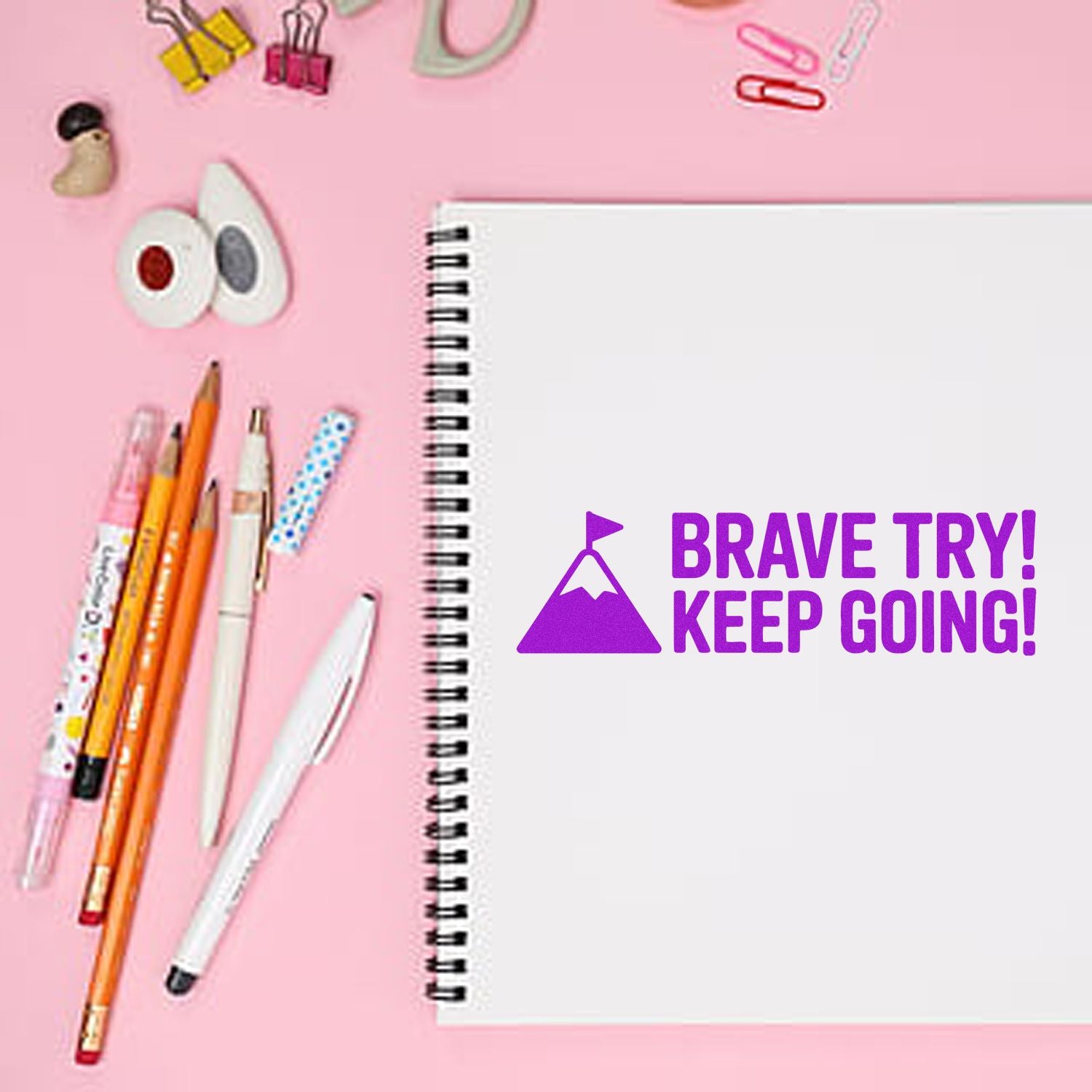 Purple motivational stamp and seal on a white spiral notebook, surrounded by pink stationery on a pastel pink background.