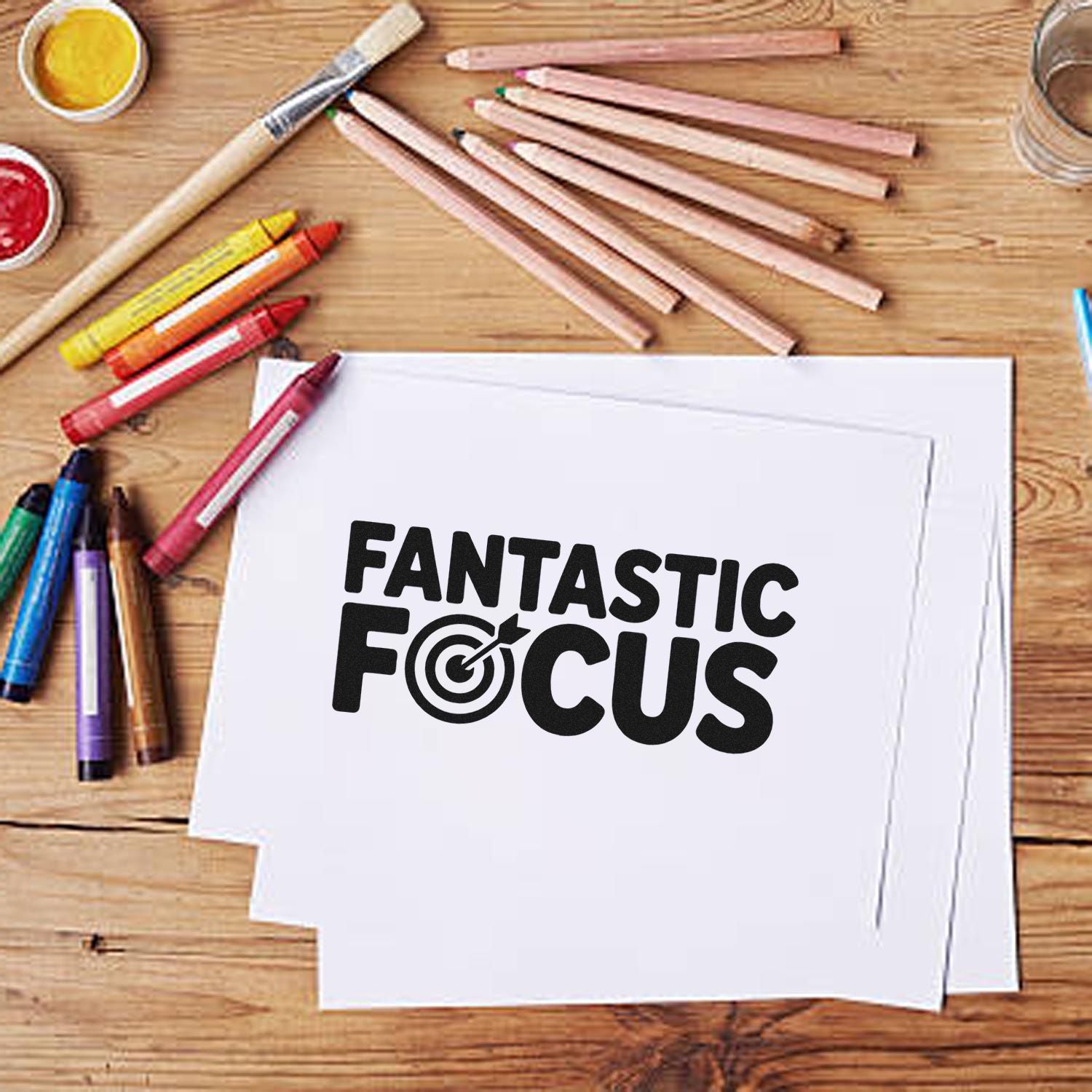 Black FANTASTIC FOCUS stamp and seal design on white paper, with colored pencils and paint on a wooden desk.