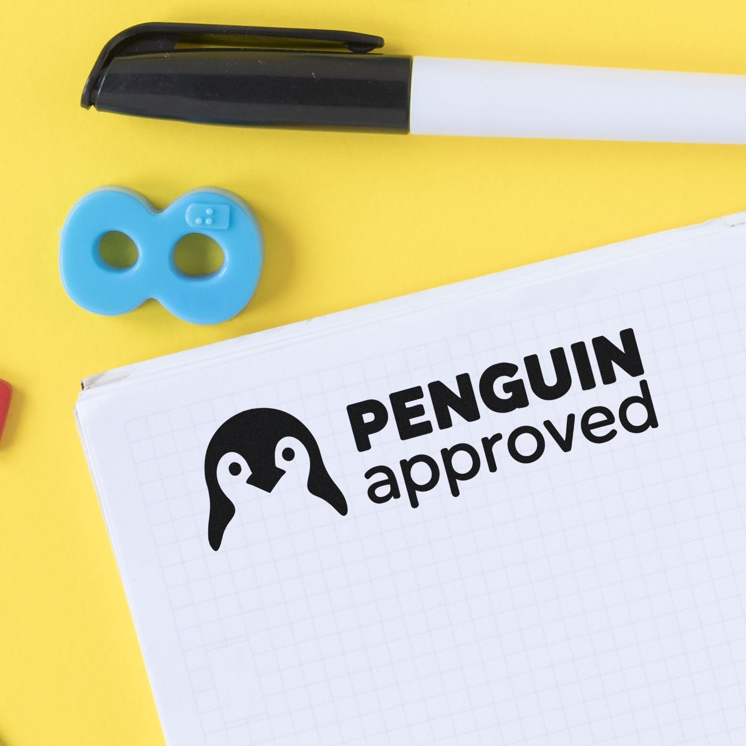 Black and white stamp pen, blue seal, and penguin approved mark on a notebook over yellow background.