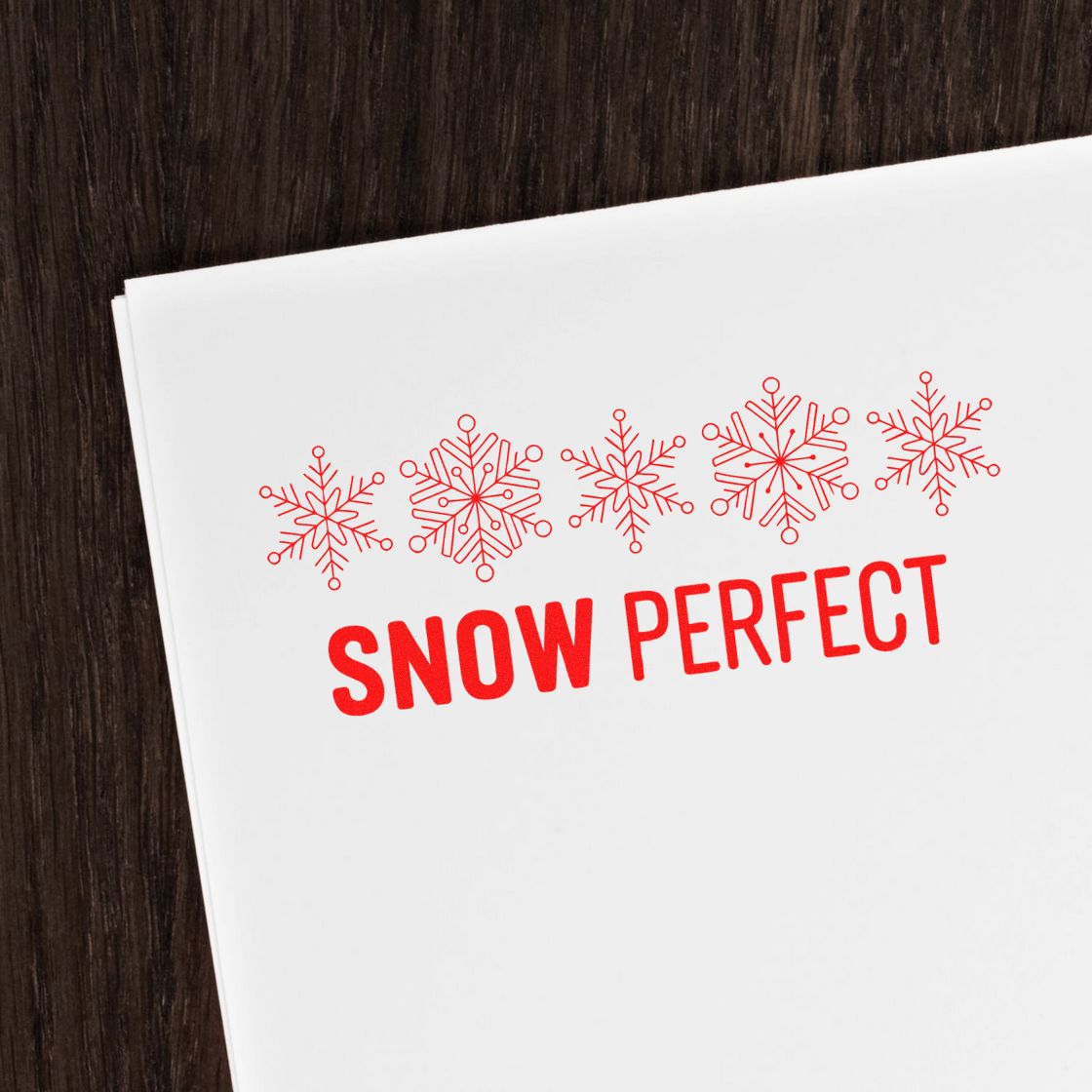 Red snowflake stamp and seal on white paper with SNOW PERFECT text, set on a dark wood background.