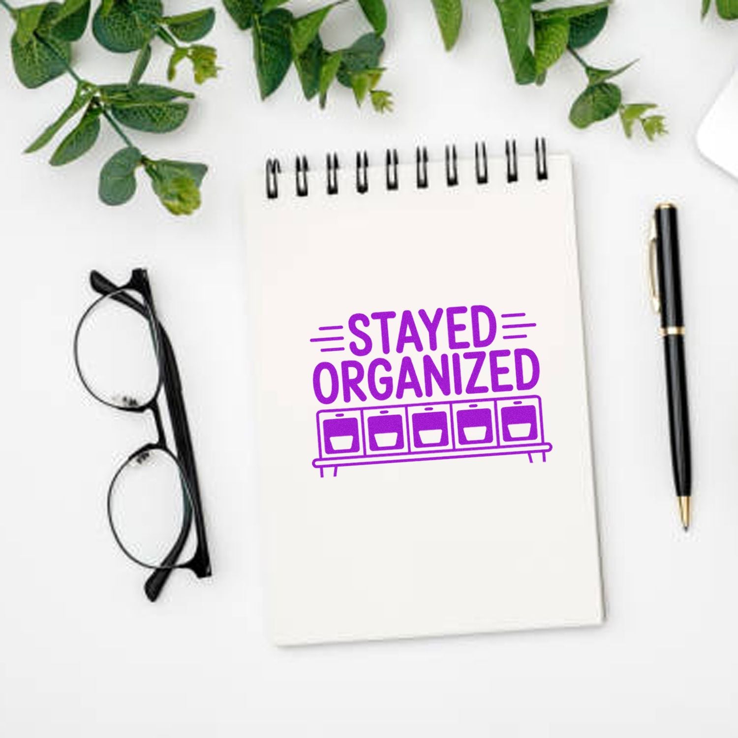 White spiral notebook with purple STAYED ORGANIZED rubber stamp seal, beside black pen and glasses on a clean desk.
