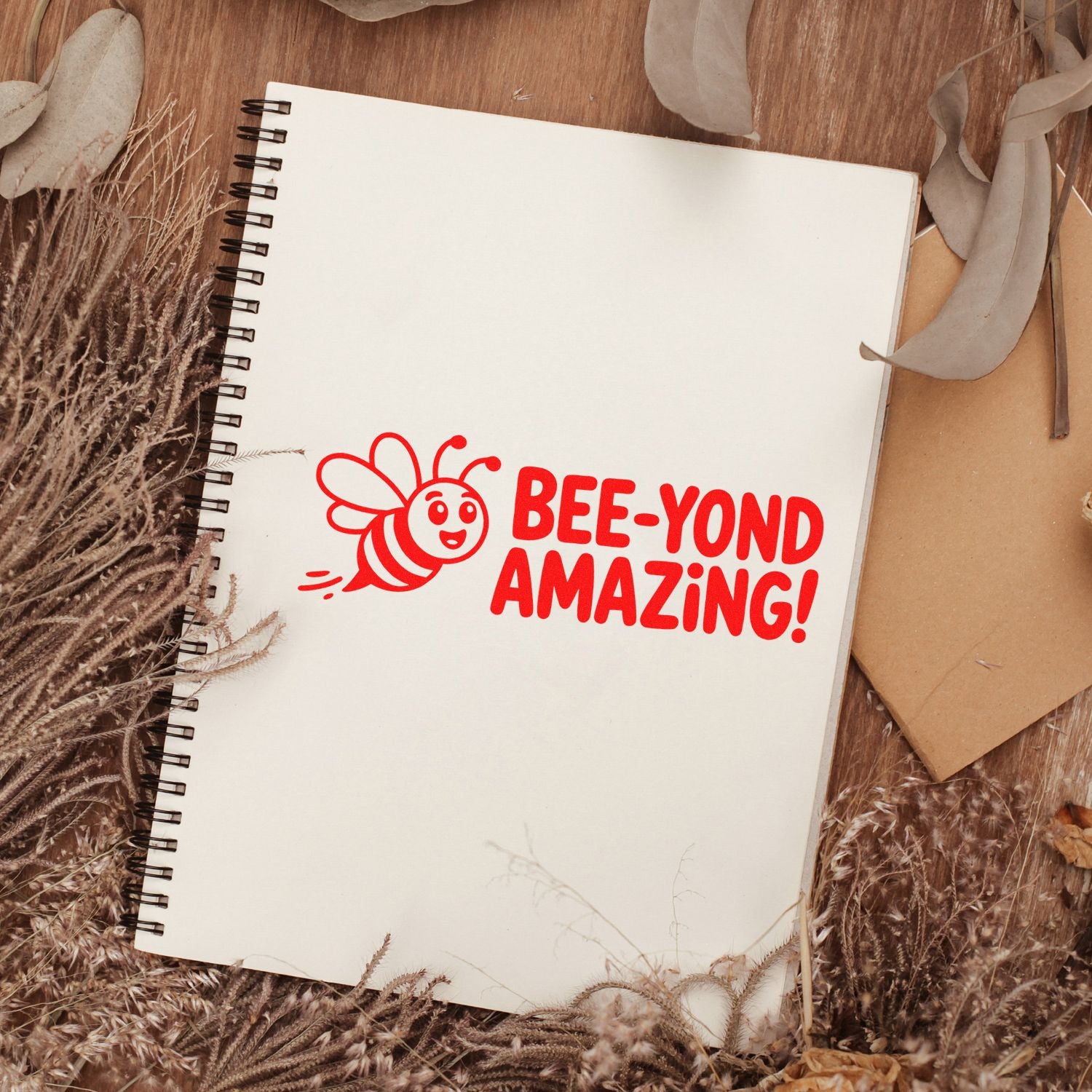 Bright red bee stamp seal reading BEE-YOND AMAZING! on a white spiral notebook with dried neutral foliage.